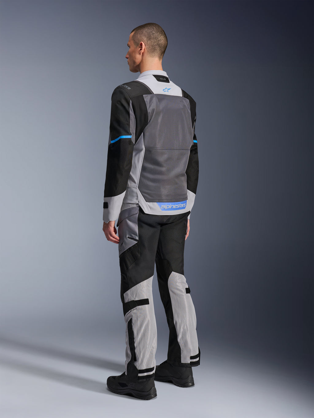 Männliches Model von hinten in der Alpinestars ST-2 Air Jacke in Ice Gray, Dark Gray und Schwarz, mit einem großen grauen Mesh-Rückeneinsatz, blauen Logo-Akzenten und passender Motorradhose für Sommertouren.