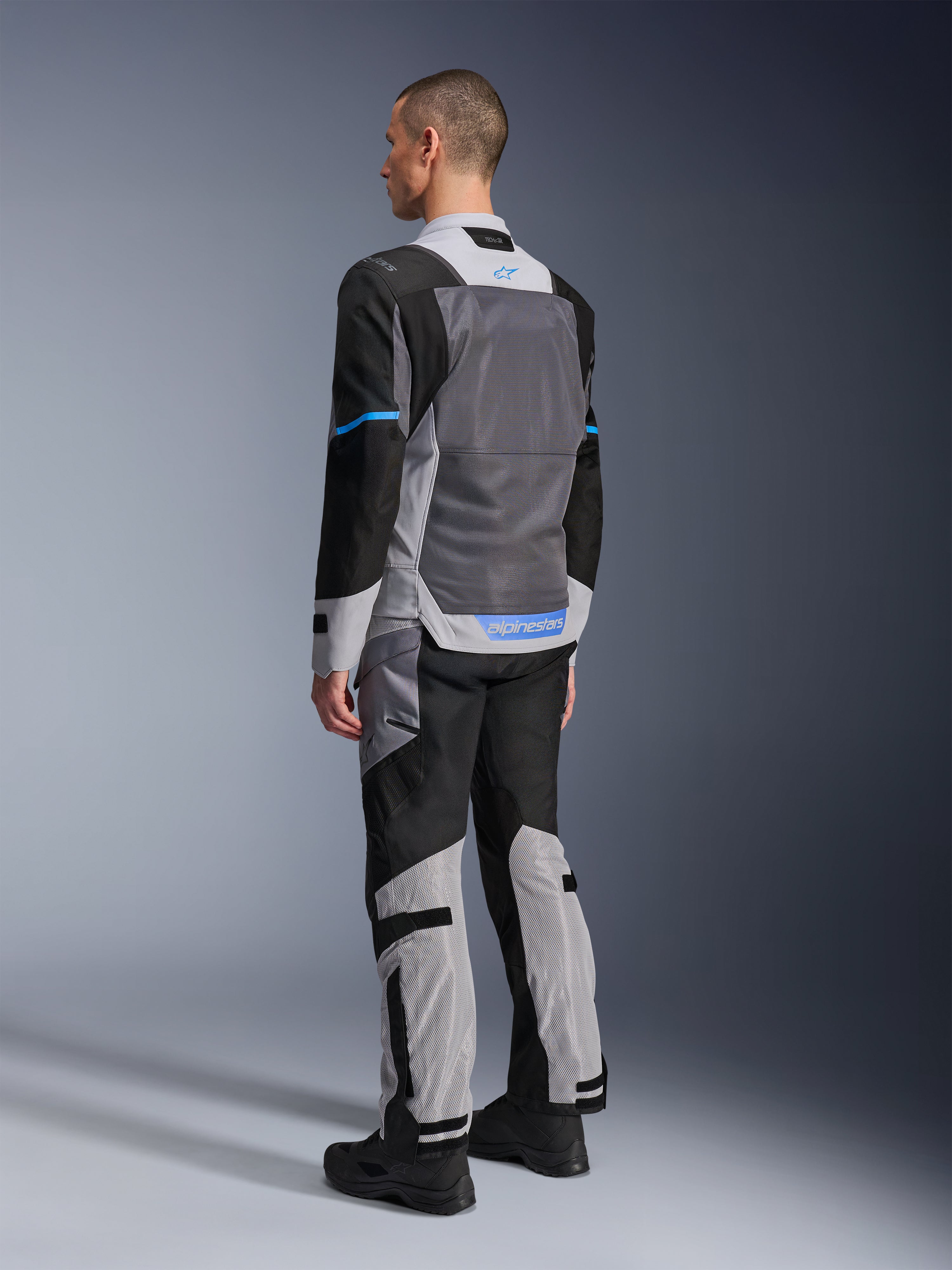 Männliches Model von hinten in der Alpinestars ST-2 Air Jacke in Ice Gray, Dark Gray und Schwarz, mit einem großen grauen Mesh-Rückeneinsatz, blauen Logo-Akzenten und passender Motorradhose für Sommertouren.