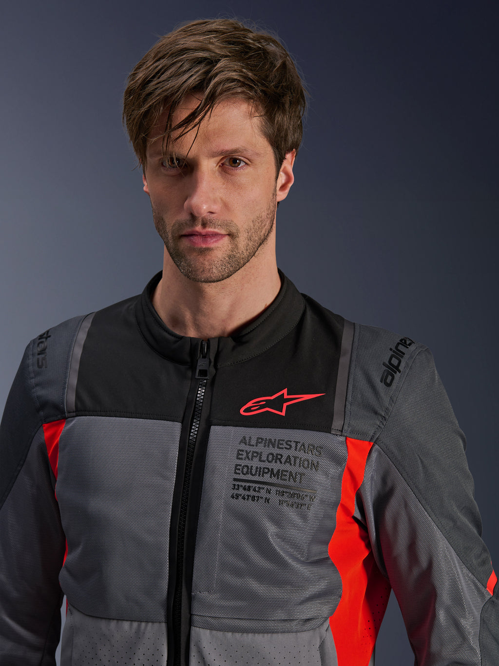 Alpinestars ST-2 Air Jacke, Deep Gray, Dark Gray und Bright Red, von einem Model getragene Sommer-Touring-Motorradjacke, mit Mesh-Belüftungseinsätzen, rotem Astars-Logo und „Exploration Equipment“-Branding auf der Brust.