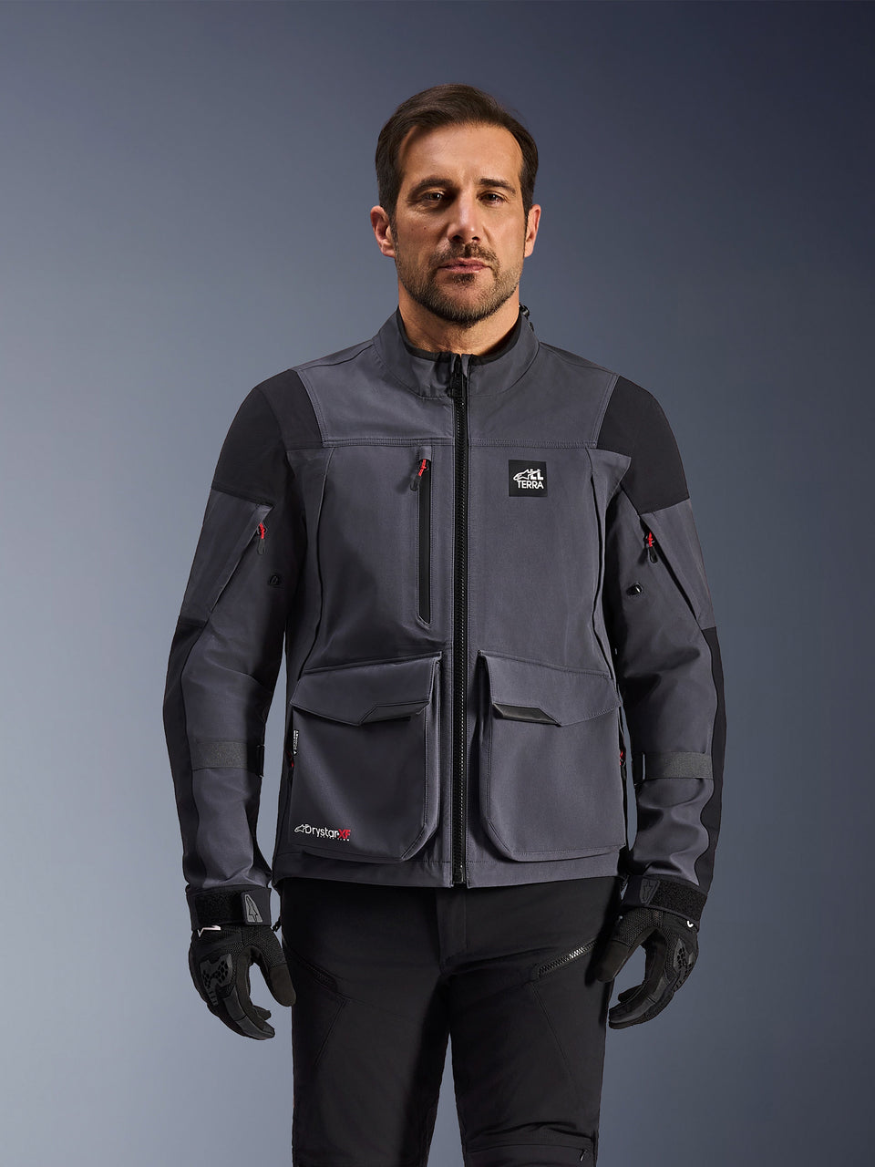 Alpinestars Maxdura Drystar®XF Laminated Armored Shell, adventure motorjas, Deep Gray en zwart, vooraanzicht op model, met grote cargozakken, 'All Terra' logo op de borst en ventilatieritsen, gedragen met zwarte motorhandschoenen en broek.
