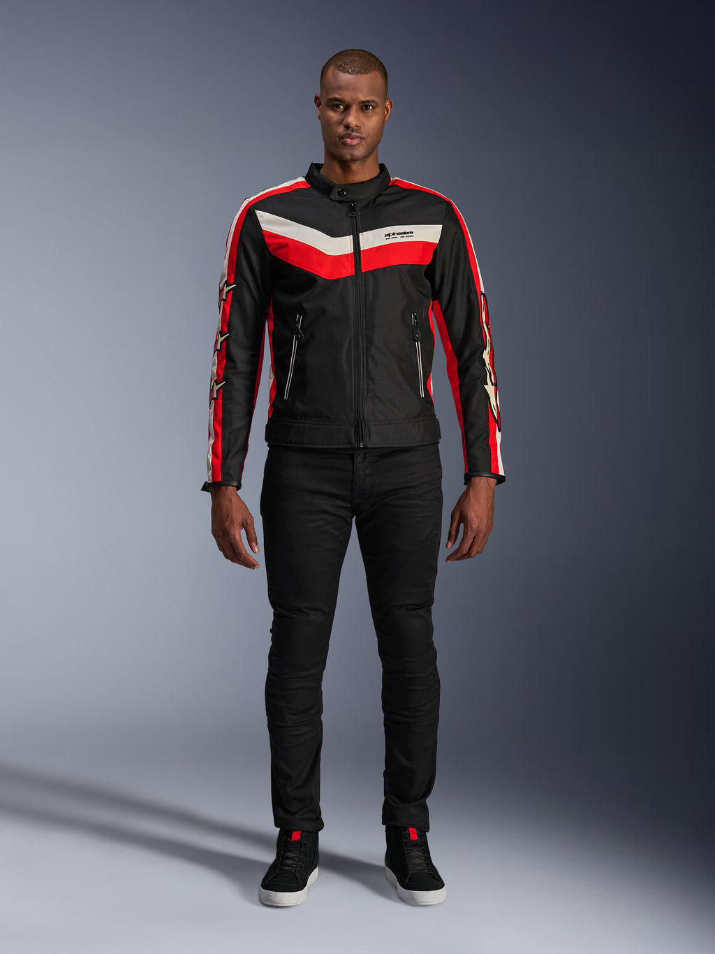Alpinestars T-Dyno WR Jacke, Textil-Motorradjacke in Schwarz, Bright Red und Ecru, mit V-förmigen Bruststreifen und Stern-Branding an den Ärmeln, getragen mit schwarzer Hose und schwarzen Motorradschuhen, Heritage-inspirierte Urban-Ausrüstung