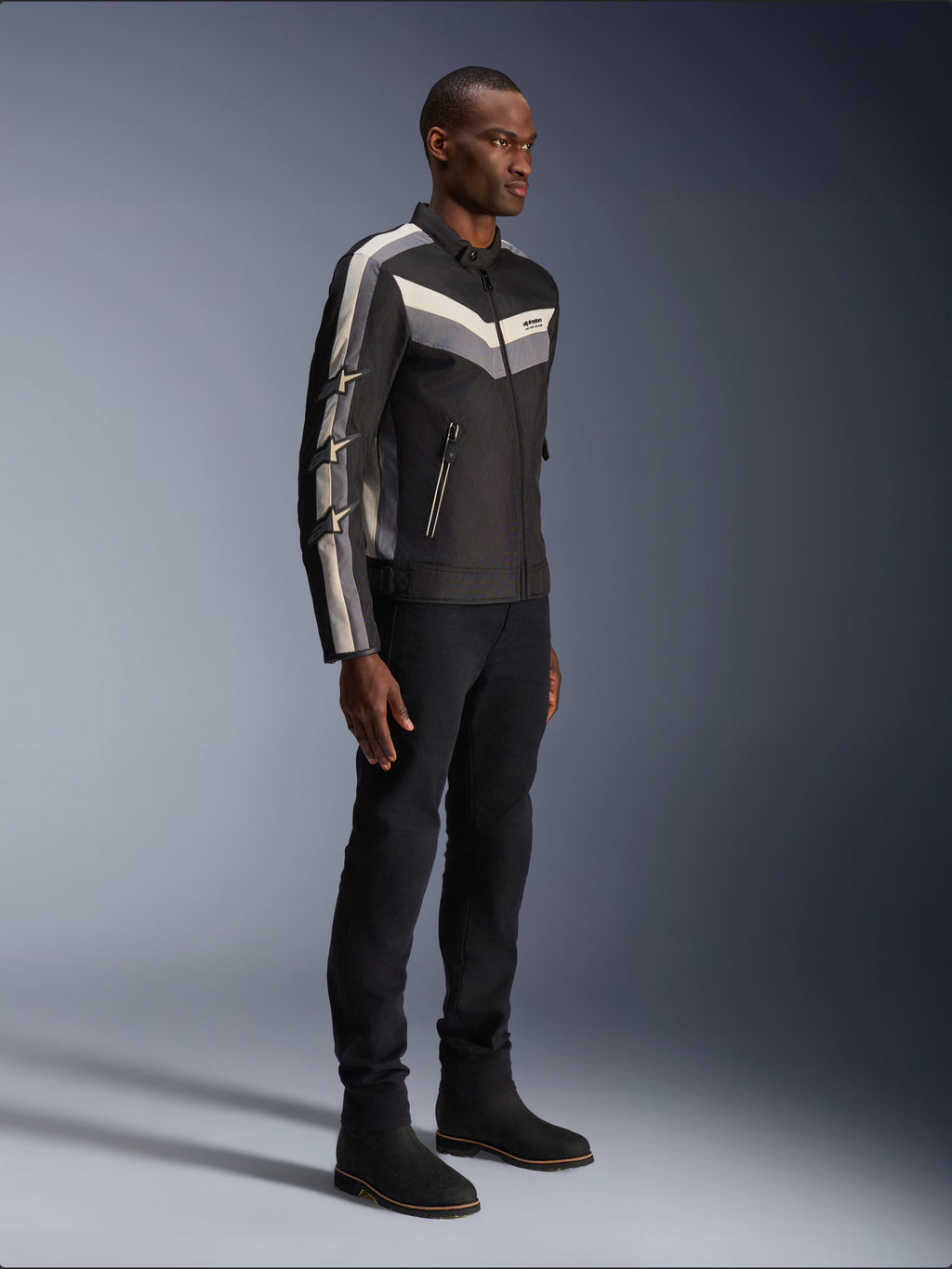 Veste Alpinestars T-Dyno WR, veste de moto en textile, noir avec accents de chevrons gris foncé et écru, coupe biker d'inspiration vintage, portée avec un pantalon et des bottes noirs pour un look routier complet