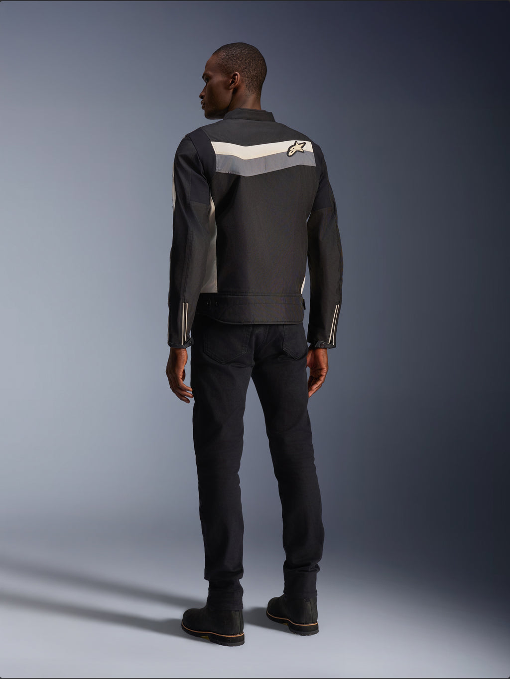 Modèle masculin de dos portant la veste Alpinestars T-Dyno WR en Noir Gris Foncé Écru, veste de moto en textile avec une bande horizontale et le logo étoile au dos, associée à un pantalon de conduite et des bottes noirs, esthétique d'équipement de moto urbain.