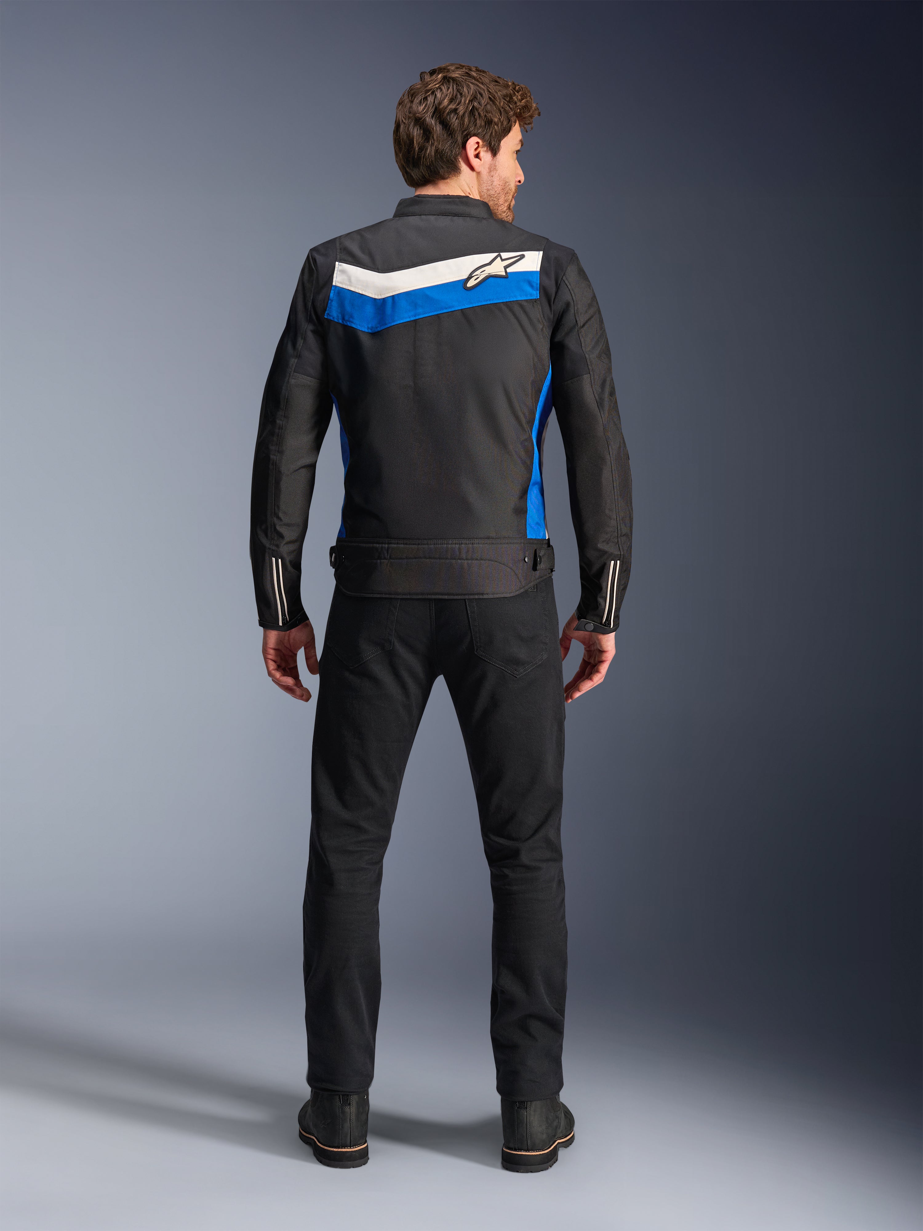 Veste Alpinestars T-Dyno WR, veste de moto en textile, coloris noir, bleu et écru, vue de dos complète sur mannequin, présentant une bande en chevron bleue et blanc cassé avec le logo Alpinestars, associée à un pantalon et des bottes noirs, équipement de conduite urbaine d'inspiration vintage