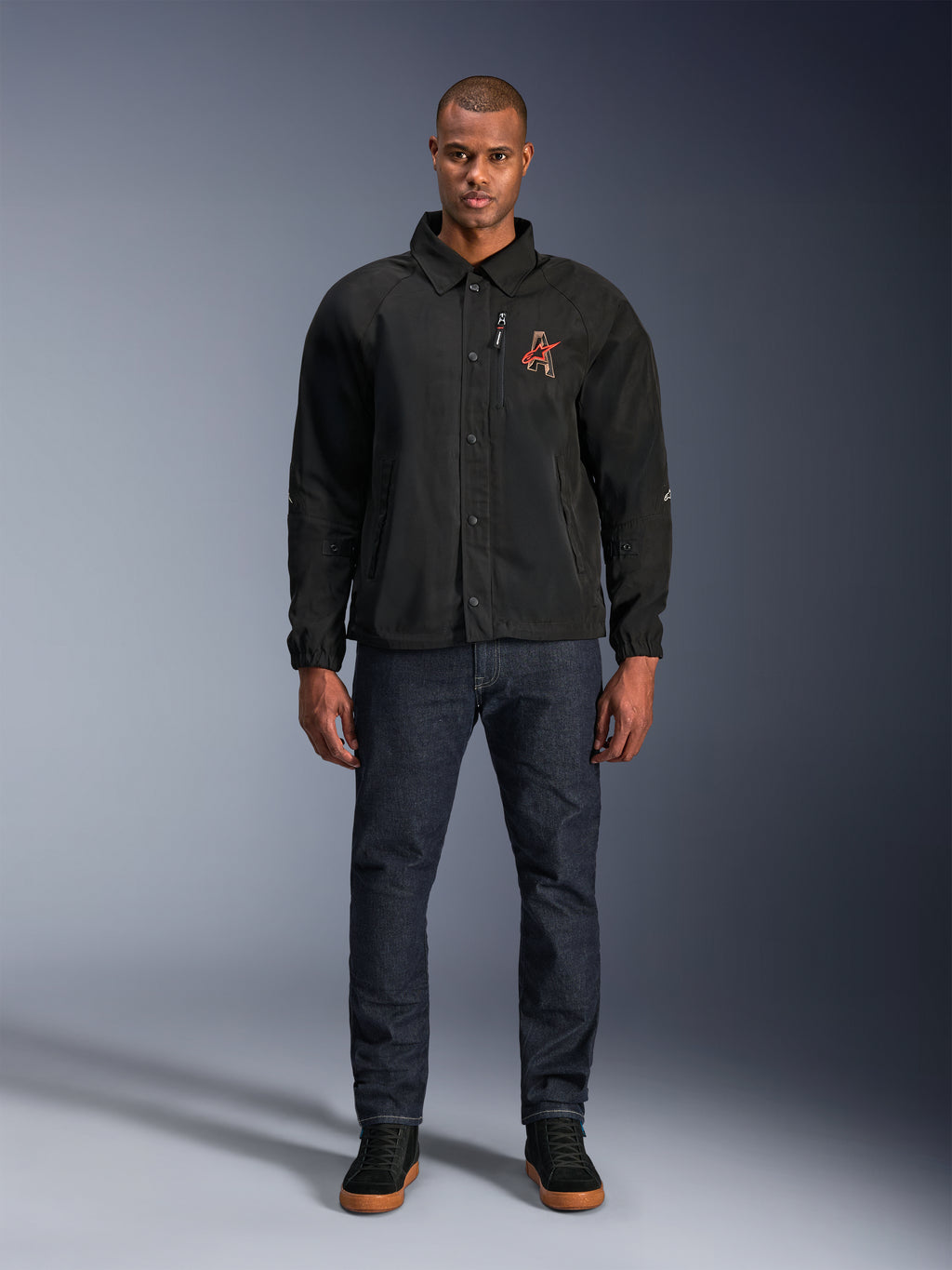 Alpinestars Revved WR Jas in het zwart, gedragen door een model met een donkerblauwe jeans en zwarte sneakers, vooraanzicht, met drukknopsluiting en een 3D geborduurd logo op de borst, urban contemporary motorkleding