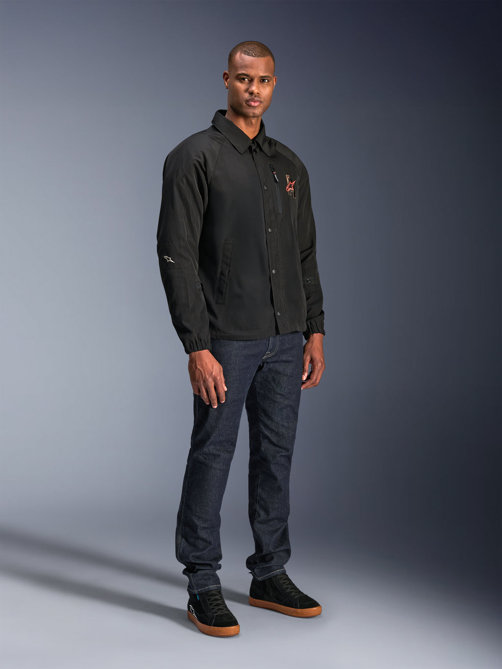 Alpinestars Revved WR Jacket in het zwart, urban motorjas in coaches-stijl gedragen door een model met donkerblauwe jeans en zwarte sneakers, voorzien van een drukknoopsluiting aan de voorzijde en een geborduurd logo, minimalistisch en sportief ontwerp voor rijden op de weg.