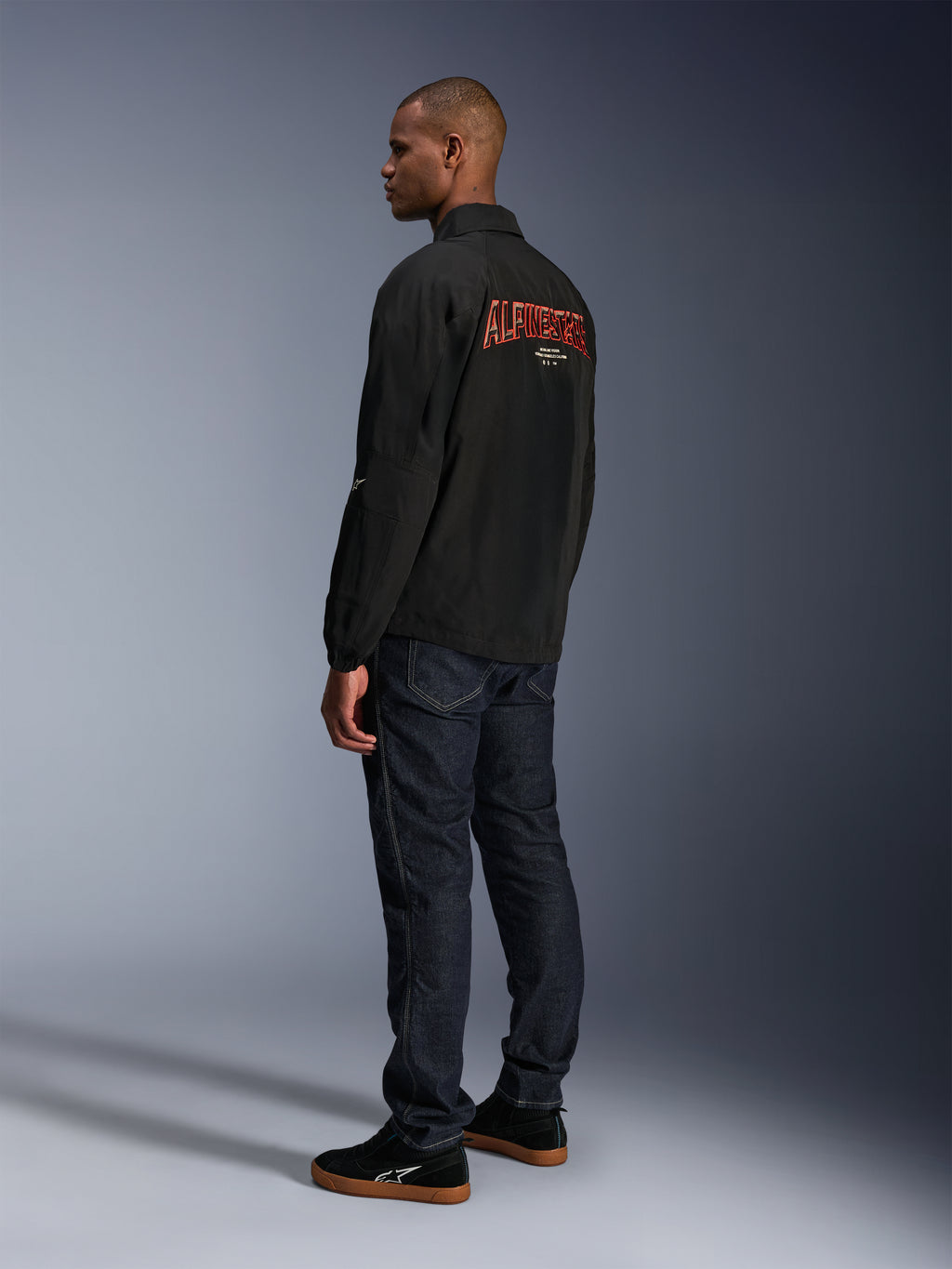 Alpinestars Revved WR Jas, zwart, met groot oranje 3D-geborduurd Alpinestars-logo op de rug, driekwart achteraanzicht van een model in donkerblauwe jeans en zwarte sneakers, urban contemporary motorjas met een coaches-jacket silhouet