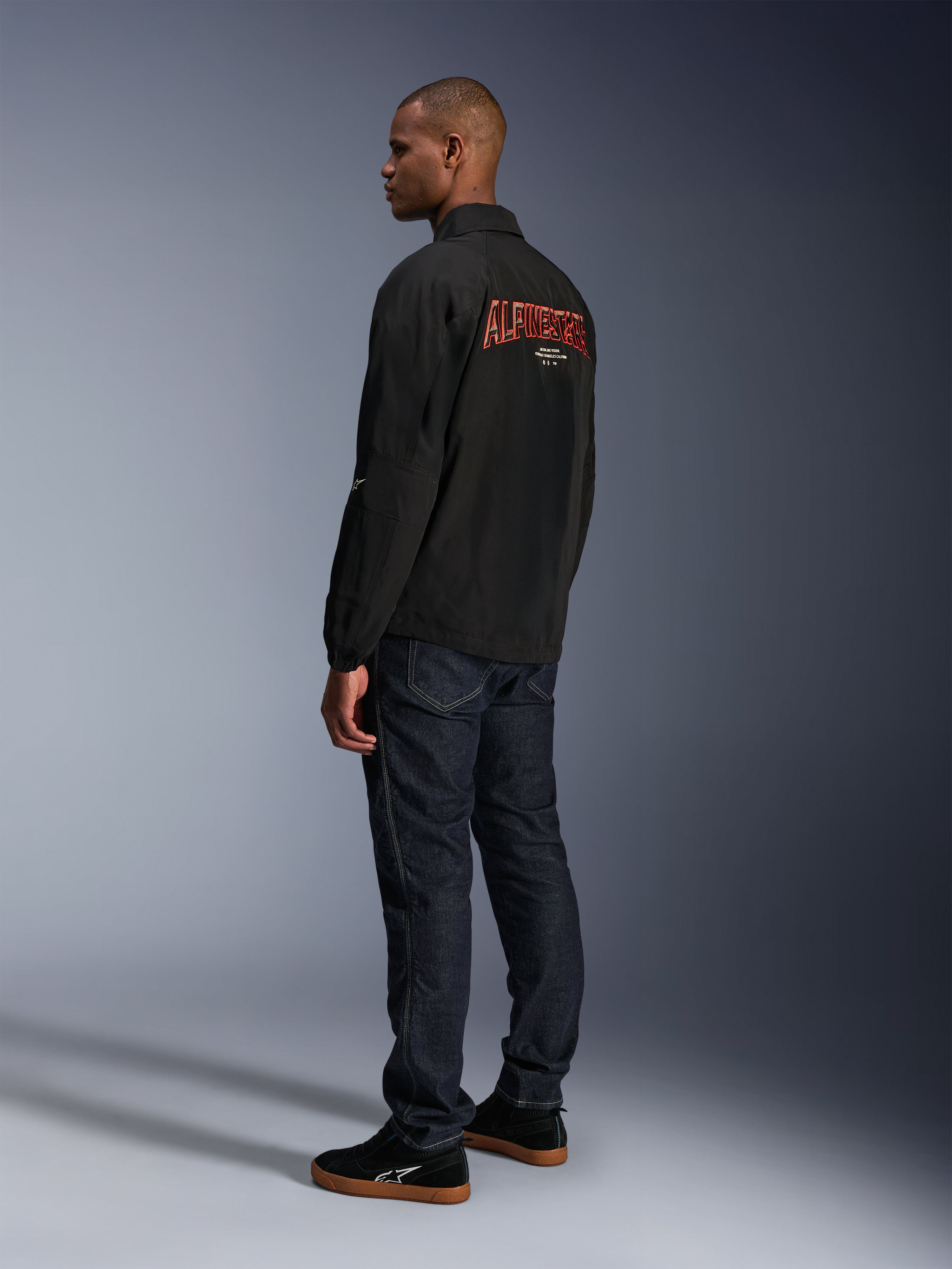 Alpinestars Revved WR Jas, zwart, met groot oranje 3D-geborduurd Alpinestars-logo op de rug, driekwart achteraanzicht van een model in donkerblauwe jeans en zwarte sneakers, urban contemporary motorjas met een coaches-jacket silhouet