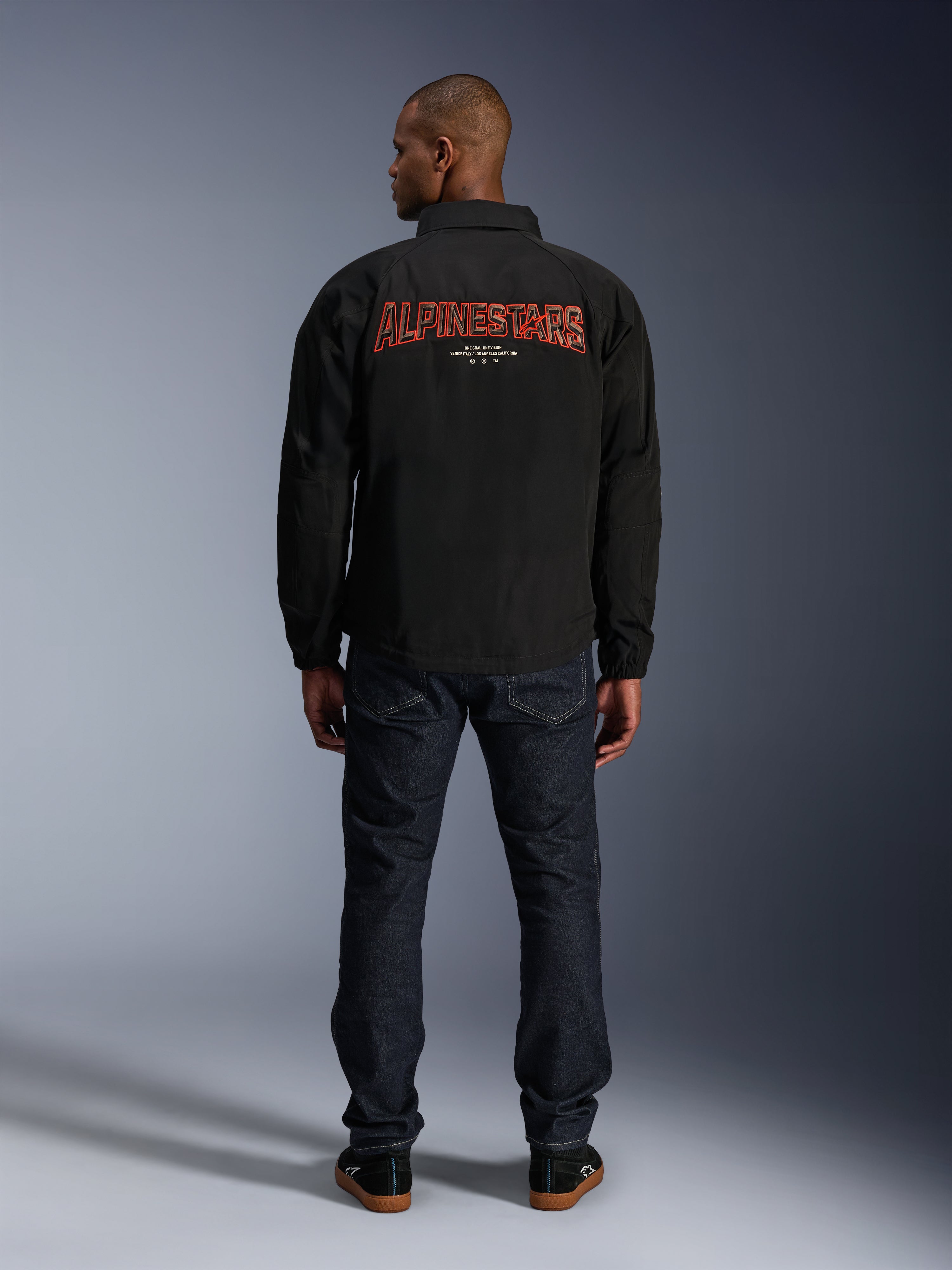 Alpinestars Revved WR Jas, zwart, achteraanzicht met groot rood 'ALPINESTARS' borduurwerk met 3D-effect, urban coachjack-stijl jas, gedragen met donkere denim jeans en zwarte sneakers, minimalistische motorkleding