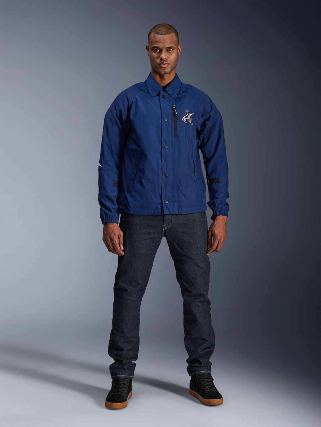 Veste Alpinestars Revved WR, bleu marine, veste de moto de style coach, portée par un homme avec un jean en denim foncé et des baskets noires, avec fermeture par boutons-pression et logo Alpinestars en 3D, streetwear urbain contemporain