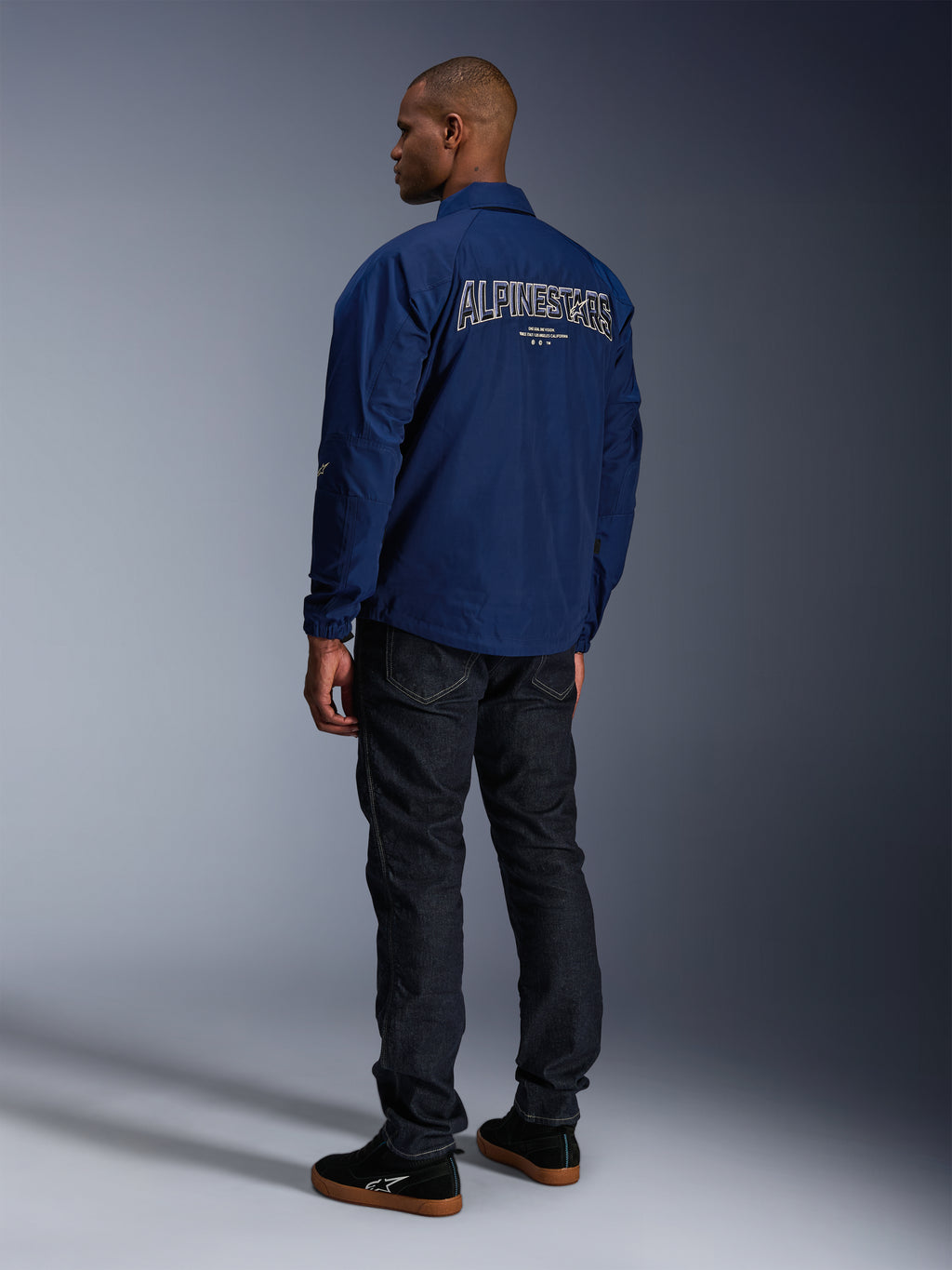 Modèle portant la veste Alpinestars Revved WR bleu marine vue de dos, avec une large broderie 'ALPINESTARS' aux contours blancs sur le dos, associée à un pantalon en denim foncé et des baskets noires, esthétique moto urbaine.