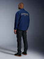 Modèle portant la veste Alpinestars Revved WR bleu marine vue de dos, avec une large broderie 'ALPINESTARS' aux contours blancs sur le dos, associée à un pantalon en denim foncé et des baskets noires, esthétique moto urbaine.