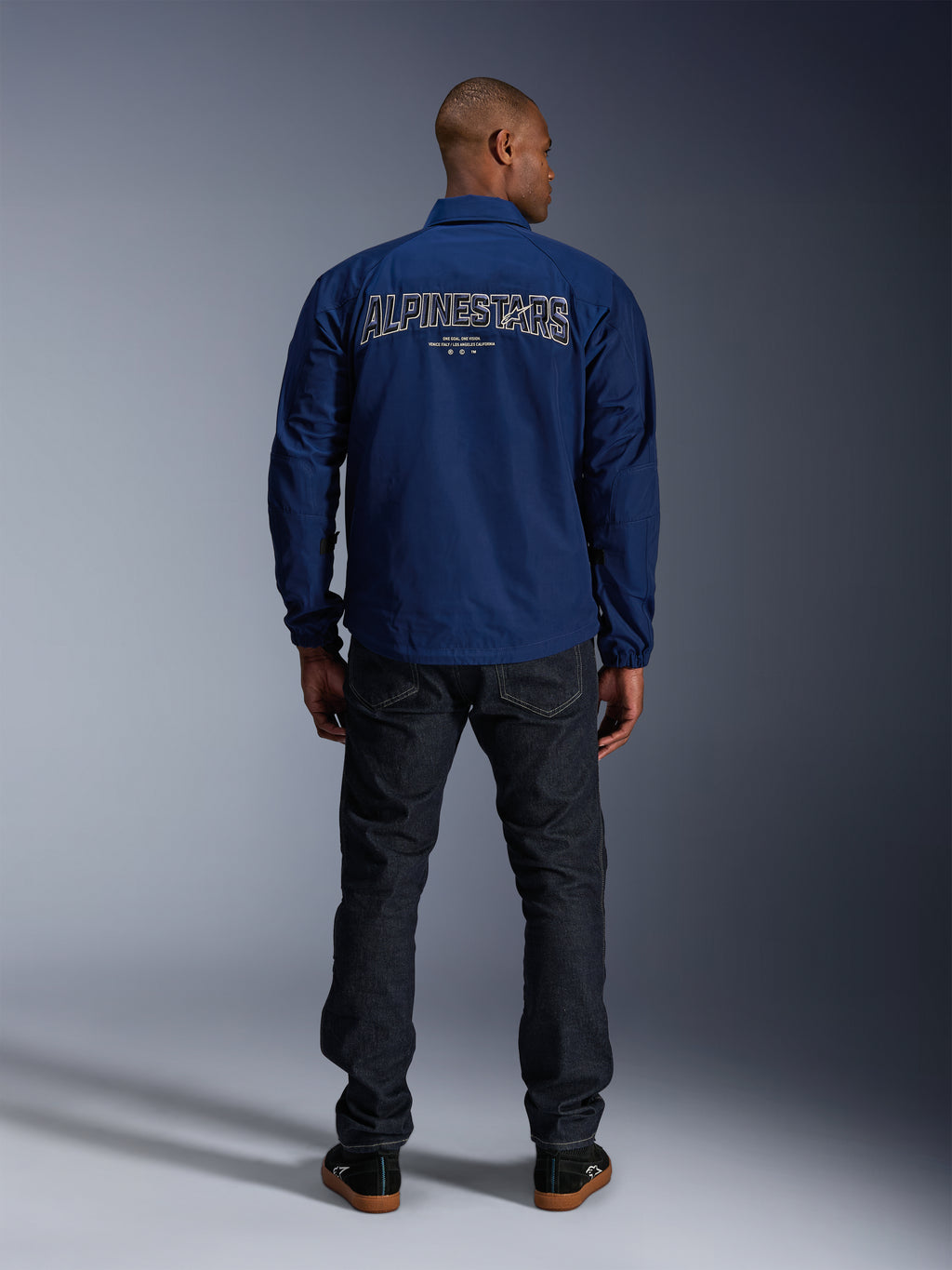 Veste Alpinestars Revved WR bleu marine, vue de dos, présentant une grande broderie 'ALPINESTARS' à effet 3D dans le dos, portée avec un jean en denim foncé et des baskets noires, vêtement de moto urbain.