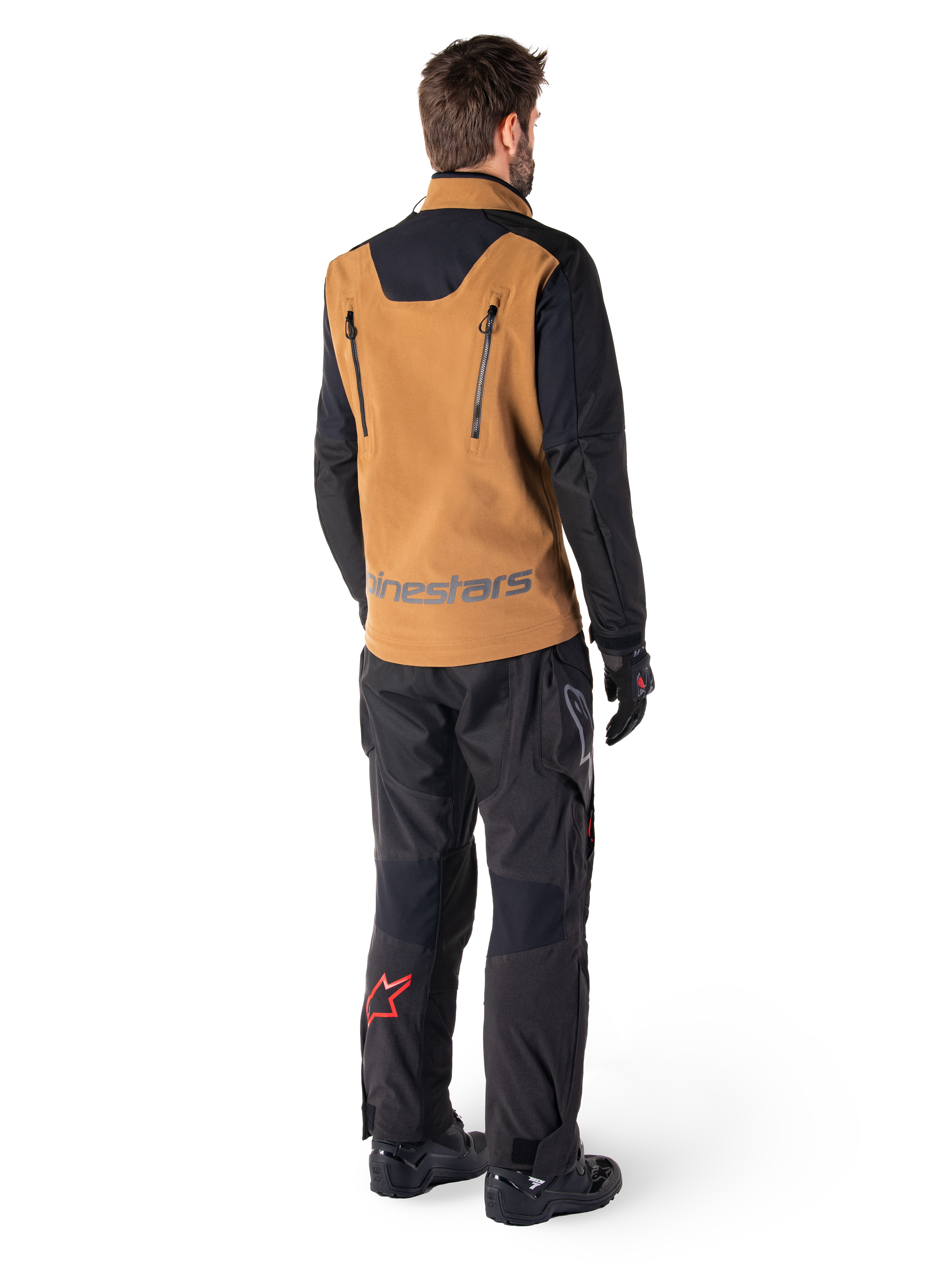 Alpinestars Hyde XT Stretch Drystar®XF Jacke und Hyde XT Drystar® Hose, Rückansicht eines Fahrers in brauner und schwarzer Adventure-Touring-Ausrüstung, Jacke mit vertikalen Reißverschlüssen am Rücken und Alpinestars-Logo, schwarze Hose mit rotem Stern-Logo an der Wade, kombiniert mit schwarzen Handschuhen und Stiefeln.