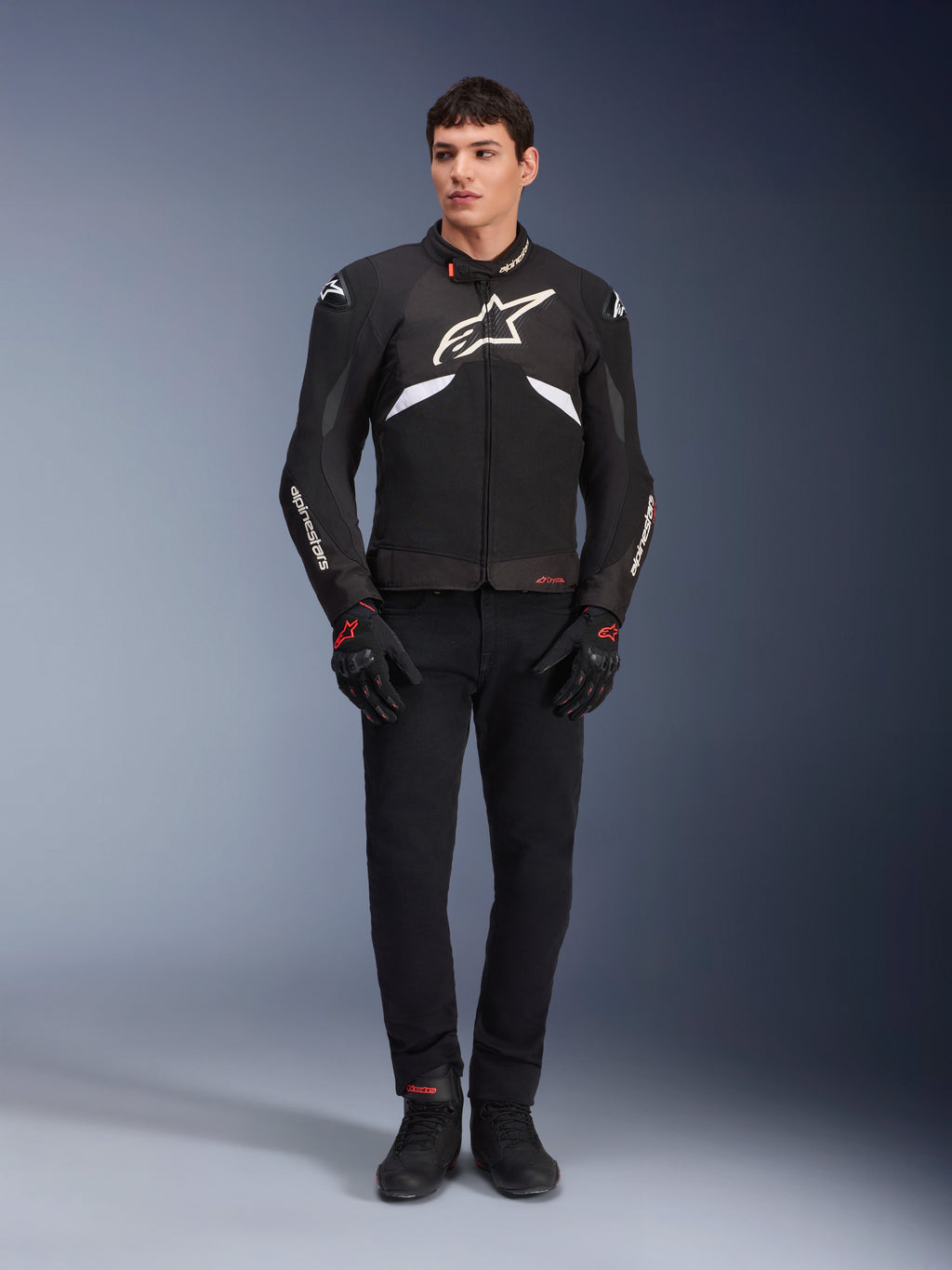 Modèle homme portant la veste Alpinestars T-GP R V3 Drystar® en noir et blanc, veste de sport textile imperméable avec un grand logo Alpinestars blanc sur la poitrine, portée avec un pantalon de moto et des gants noirs, portrait professionnel en studio soulignant la silhouette sportive élégante.