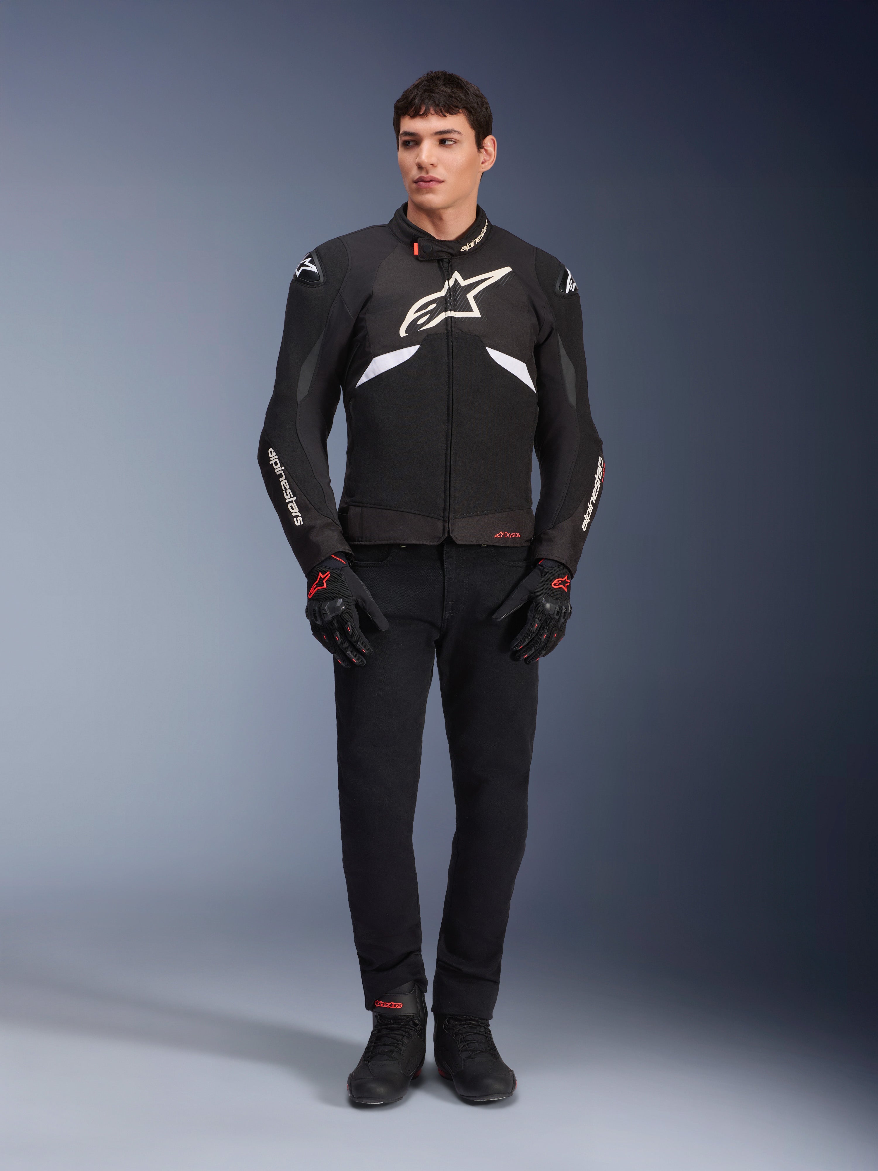 Modèle homme portant la veste Alpinestars T-GP R V3 Drystar® en noir et blanc, veste de sport textile imperméable avec un grand logo Alpinestars blanc sur la poitrine, portée avec un pantalon de moto et des gants noirs, portrait professionnel en studio soulignant la silhouette sportive élégante.