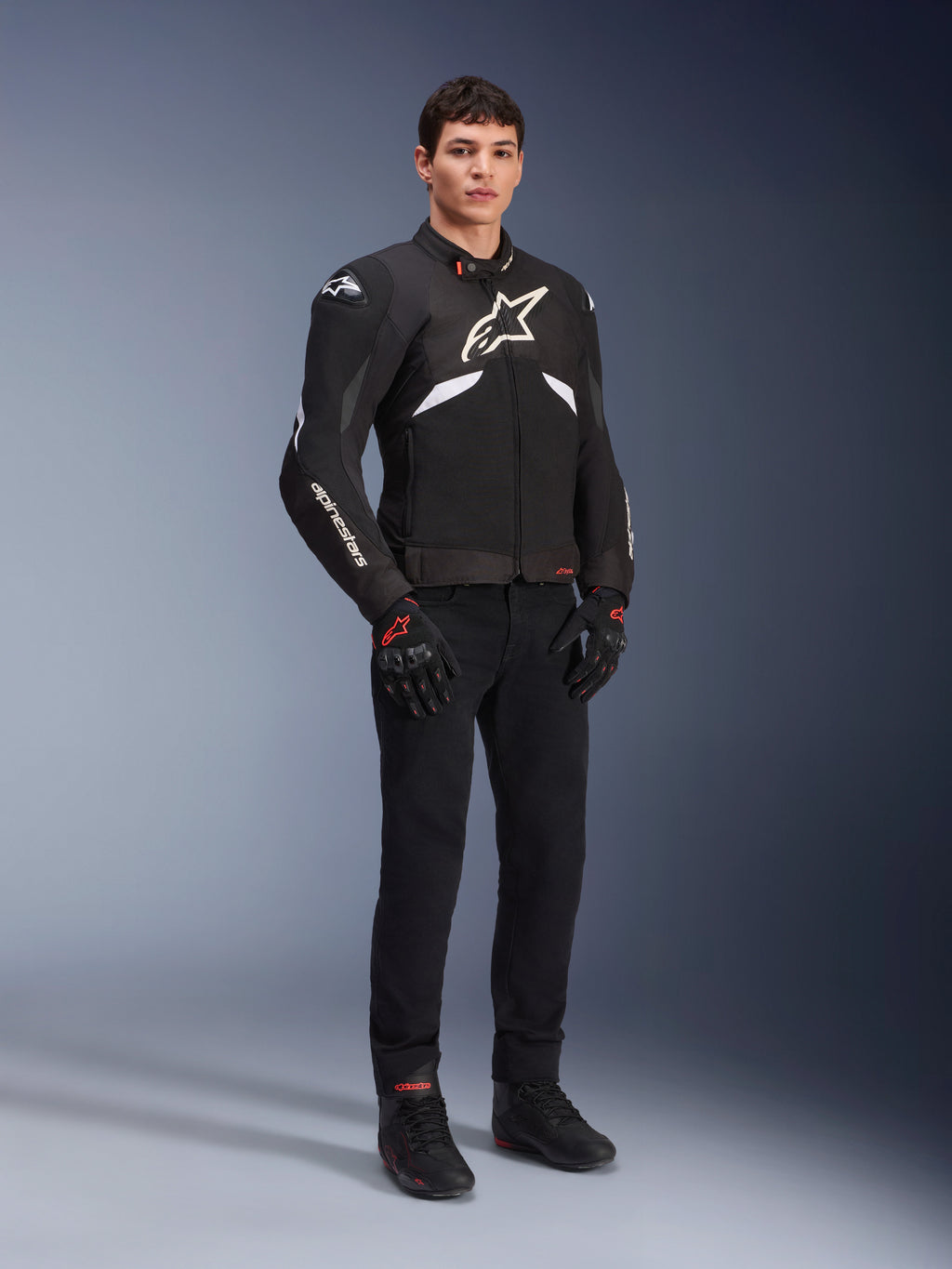 Veste Alpinestars T-GP R V3 Drystar® en noir et blanc, portée par un mannequin avec gants et pantalon de moto noirs, présentant un logo blanc sur la poitrine, des touches de blanc sur les manches et des sliders de protection aux épaules, veste de sport textile imperméable pour la route