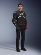 Veste Alpinestars T-GP R V3 Drystar® en noir et blanc, portée par un mannequin avec gants et pantalon de moto noirs, présentant un logo blanc sur la poitrine, des touches de blanc sur les manches et des sliders de protection aux épaules, veste de sport textile imperméable pour la route