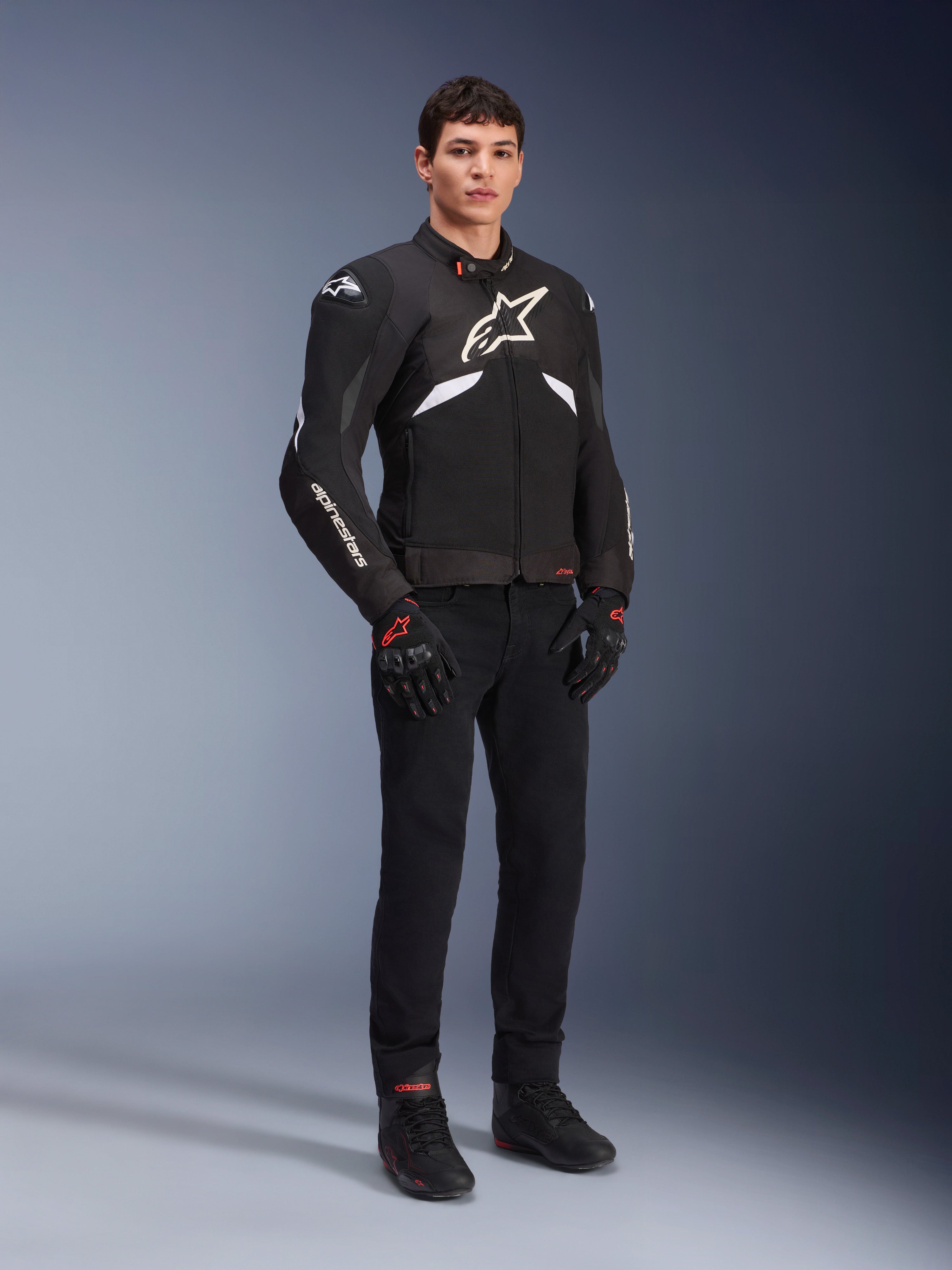 Veste Alpinestars T-GP R V3 Drystar® en noir et blanc, portée par un mannequin avec gants et pantalon de moto noirs, présentant un logo blanc sur la poitrine, des touches de blanc sur les manches et des sliders de protection aux épaules, veste de sport textile imperméable pour la route
