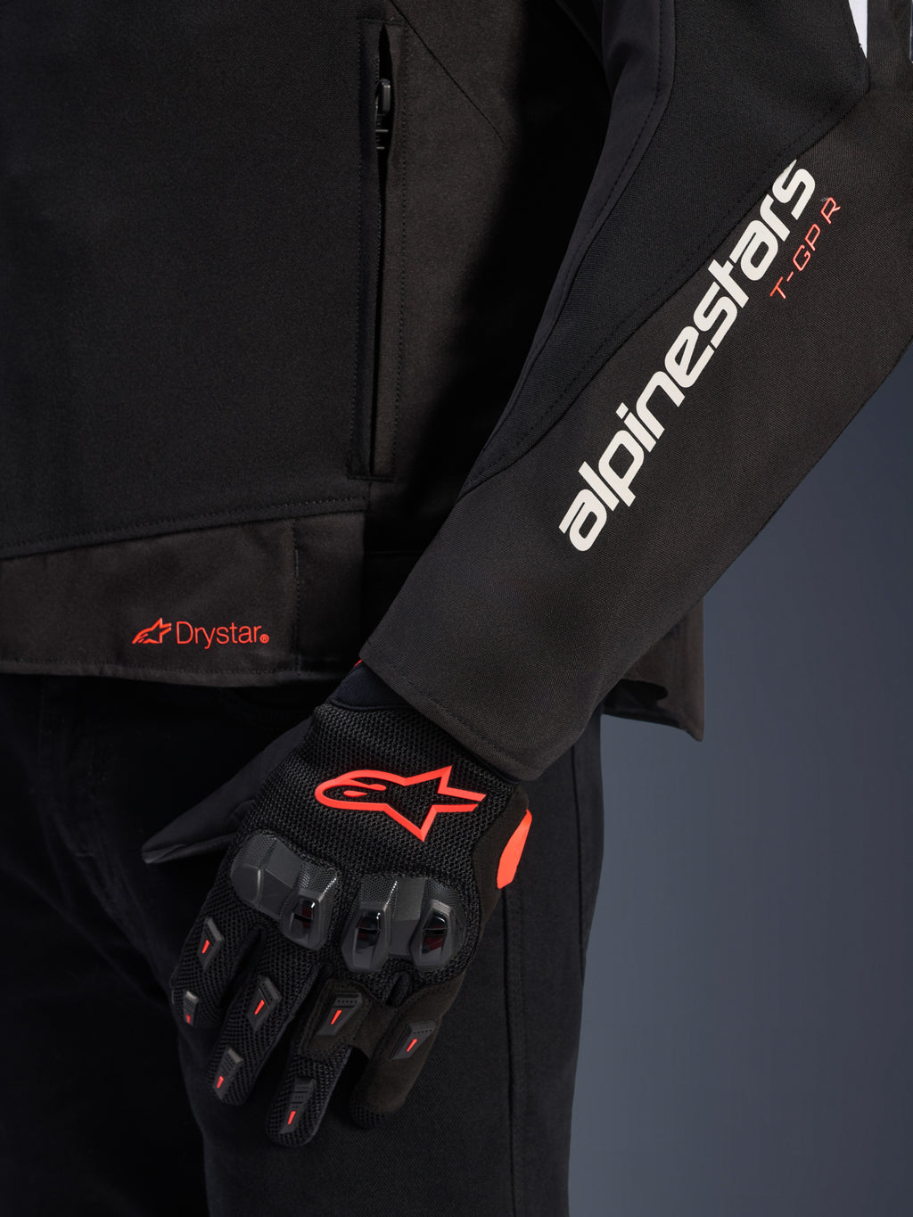 Veste Alpinestars T-GP R V3 Drystar®, veste de moto en textile noir, gros plan montrant le marquage blanc Alpinestars et orange T-GP R sur la manche, logo Drystar orange sur l'ourlet, associée à des gants de protection en mesh noir avec des accents rouges.