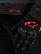 Gros-plan sur la taille de la veste Alpinestars T-GP R V3 Drystar® avec un logo Drystar rouge, accompagnée d'un gant de moto noir avec logo étoile Alpinestars rouge, tissu mesh et protection des articulations renforcée.