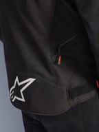 Veste Alpinestars T-GP R V3 Drystar®, noir et blanc, vue rapprochée du bas du dos présentant un grand logo étoile Alpinestars blanc, une poche latérale zippée avec tirette rouge et une sangle de réglage velcro à la taille, matériau textile durable.