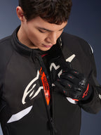 Veste Alpinestars T-GP R V3 Drystar®, veste textile sport moto noir et blanc, partiellement ouverte révélant la sangle intérieure orange « Alpinestars Protects », portée avec des gants en mesh noir et rouge, équipement de protection dérivé de la course.