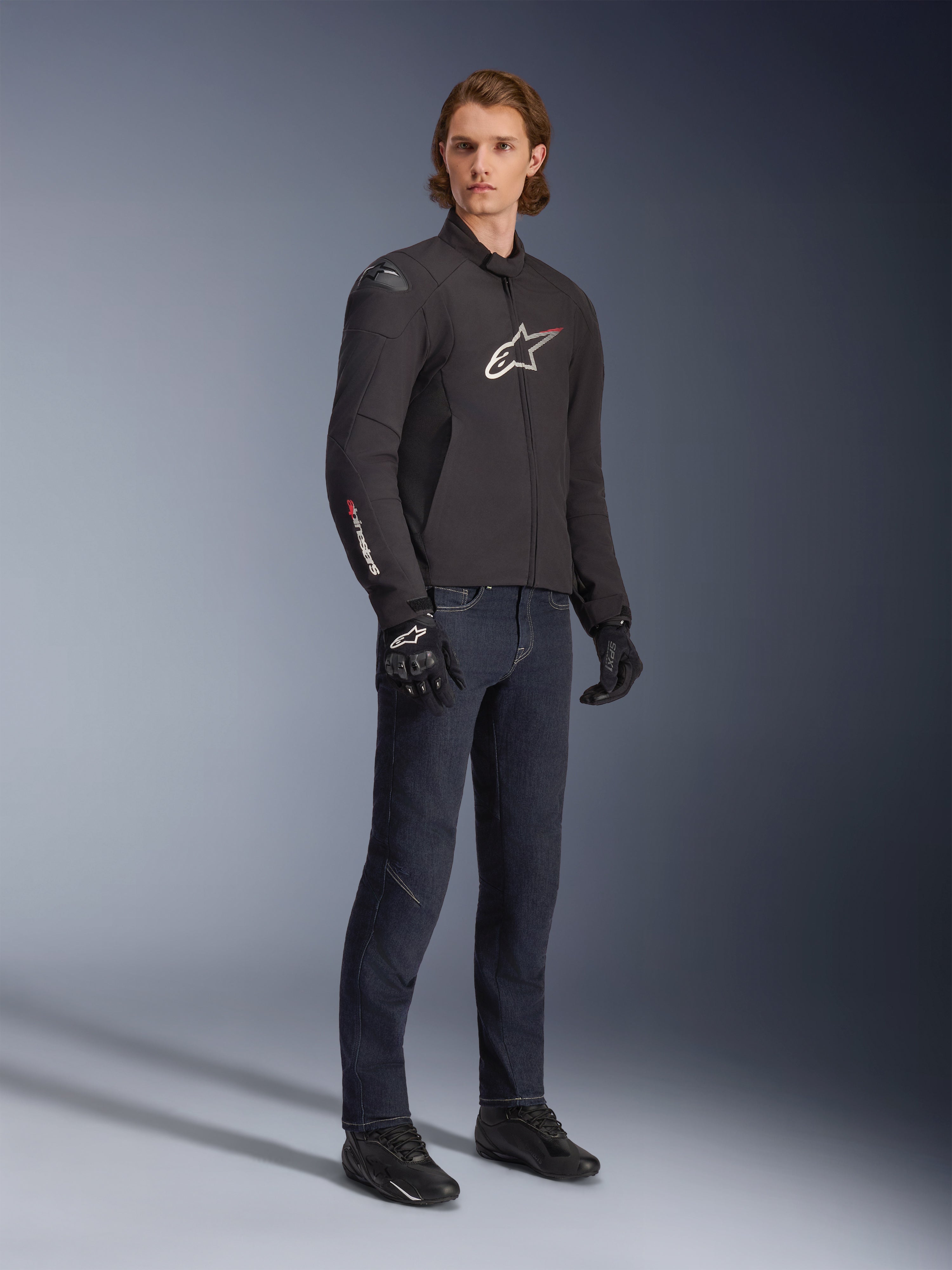 Männliches Model trägt Alpinestars SMX Waterproof Jacke, Schwarz mit weißen und roten Logo-Akzenten, kombiniert mit dunkelblauer Denim-Hose und schwarzen Motorradhandschuhen, schlanke Textilmotorradjacke mit sportlicher Passform.