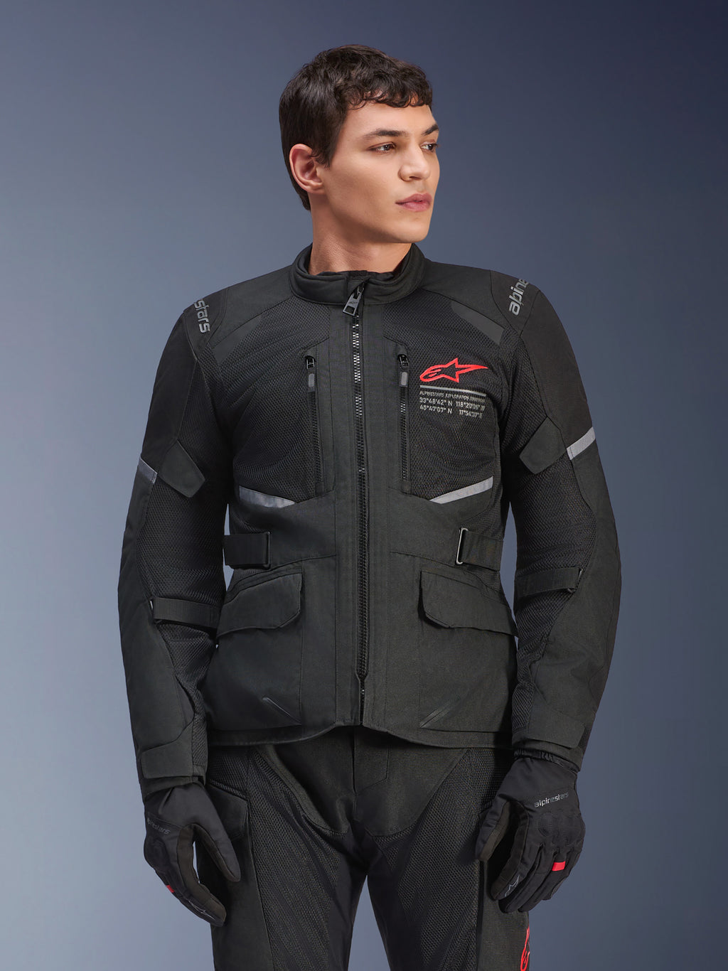 Alpinestars Andes Air Drystar® jas, zwarte touring motorjas, rood sterlogo op de borst, voorzien van mesh-ventilatiepanelen en reflecterende accenten, gedragen door mannelijk model met zwarte motorhandschoenen, adventure-uitrusting voor twee seizoenen