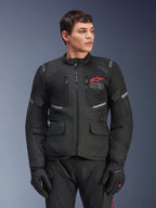 Alpinestars Andes Air Drystar® jas, zwarte touring motorjas, rood sterlogo op de borst, voorzien van mesh-ventilatiepanelen en reflecterende accenten, gedragen door mannelijk model met zwarte motorhandschoenen, adventure-uitrusting voor twee seizoenen