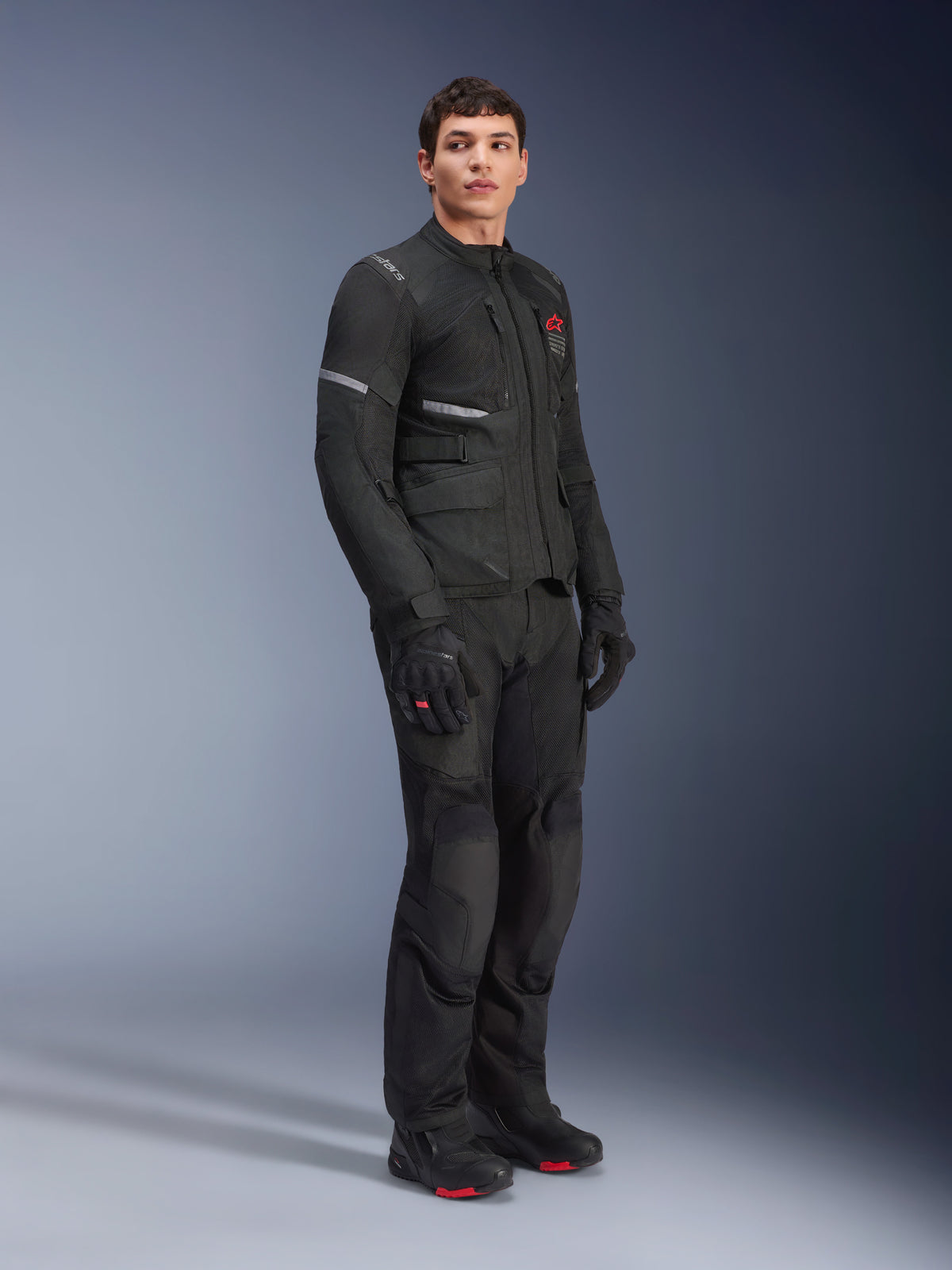 Alpinestars Andes Air Drystar® Jas, zwart, gedragen door een model als onderdeel van een volledige motoradventure-uitrusting met bijpassende broek, handschoenen en laarzen, voorzien van mesh ventilatiepanelen, een rood logo op de borst en reflecterende strips voor touring-zichtbaarheid.