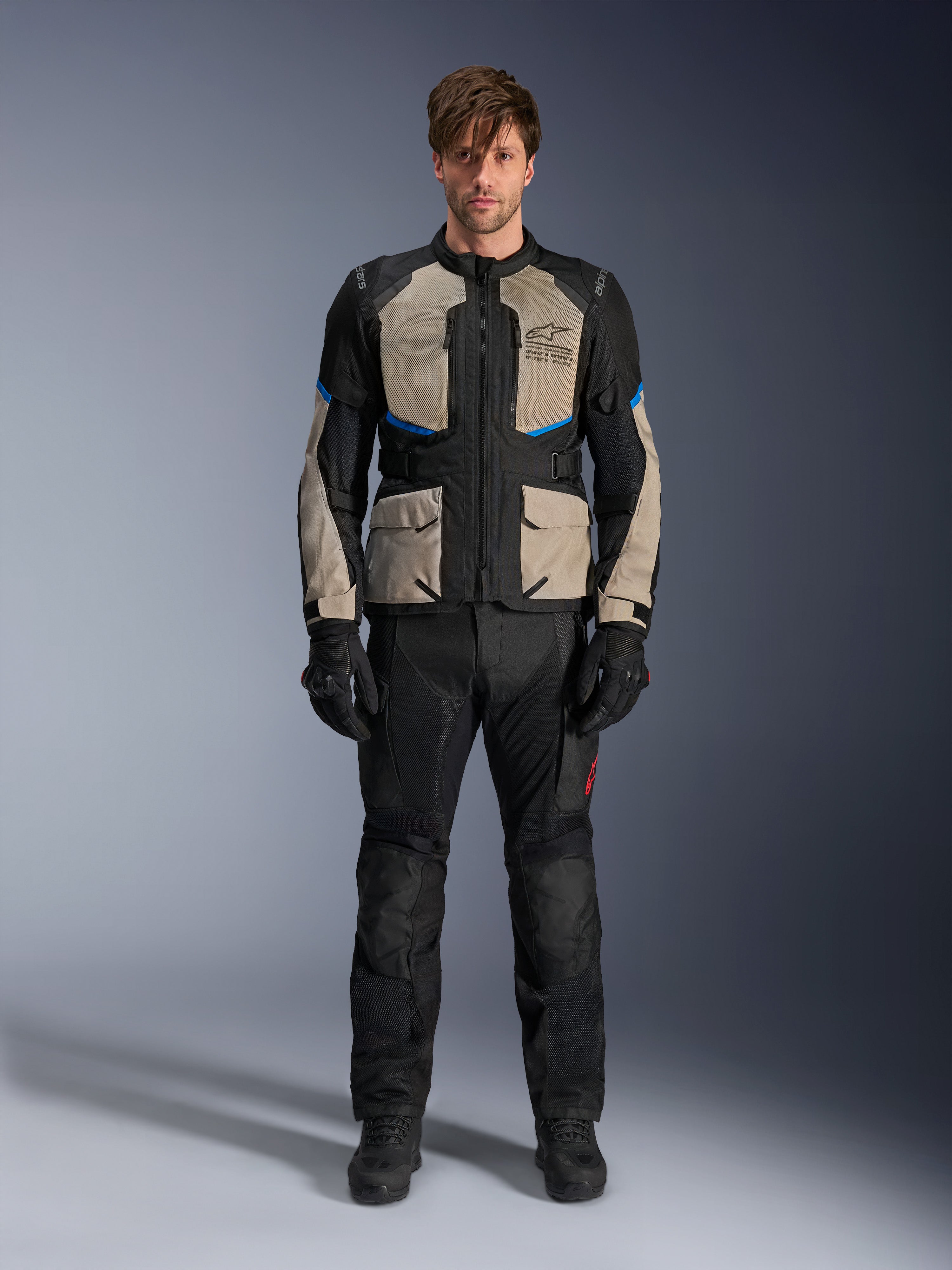 Modell trägt Alpinestars Andes Air Drystar® Jacke, Motorrad-Tourenjacke, Schwarz, Laurel Gray und Bright Blue, kombiniert mit passender schwarzer Fahrhose und Handschuhen, mit großflächigen Mesh-Belüftungseinsätzen an Brust und Ärmeln für Adventure-Touren bei warmem Wetter.