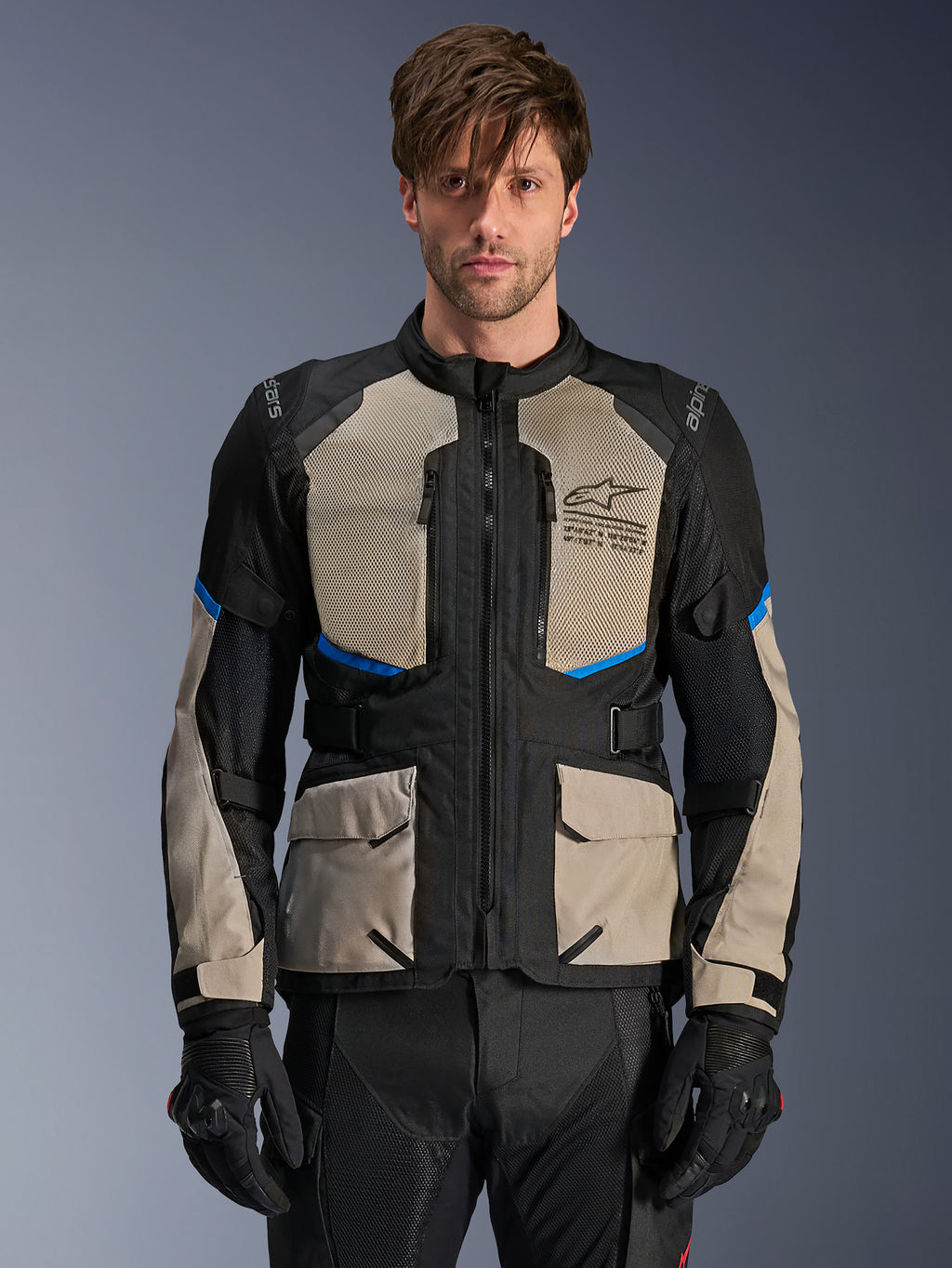 Alpinestars Andes Air Drystar Jacke, Adventure-Touringjacke, Schwarz, Laurel Gray und Bright Blue, getragen von einem Modell mit schwarzen Handschuhen, mit großen Mesh-Einsätzen im Brustbereich für Luftzirkulation, vorderen Cargotaschen und blauen Akzenten, schützende Motorrad-Tourenausrüstung