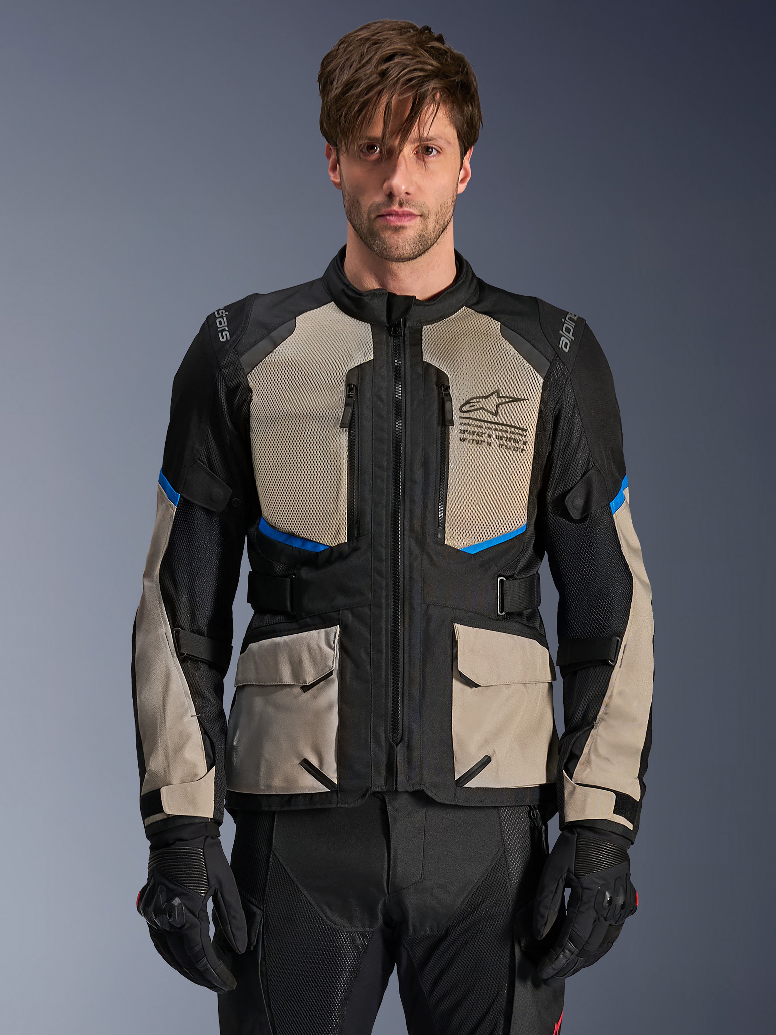 Alpinestars Andes Air Drystar motorjas, adventure touringjas, zwart, Laurel grijs en helderblauw, gedragen door een model met zwarte handschoenen, voorzien van grote mesh borstpanelen voor luchtstroom, cargo-zakken aan de voorzijde en blauwe accenten, beschermende motorkleding