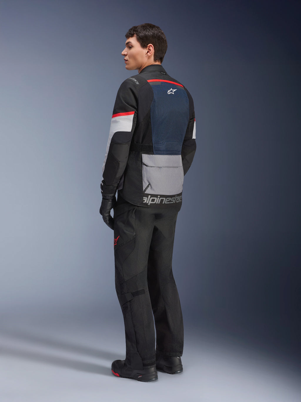 Männliches Model in der Rückansicht von der Seite, das die Alpinestars Andes Air Drystar® Jacke in Deep Blue Black Ice Gray trägt, kombiniert mit schwarzer Motorradhose und Handschuhen, mit blauem Mesh-Rückeneinsatz, grauer Utility-Tasche am unteren Rücken und roten Akzenten an den Ärmeln, professionelle Adventure-Touring-Ausrüstung.
