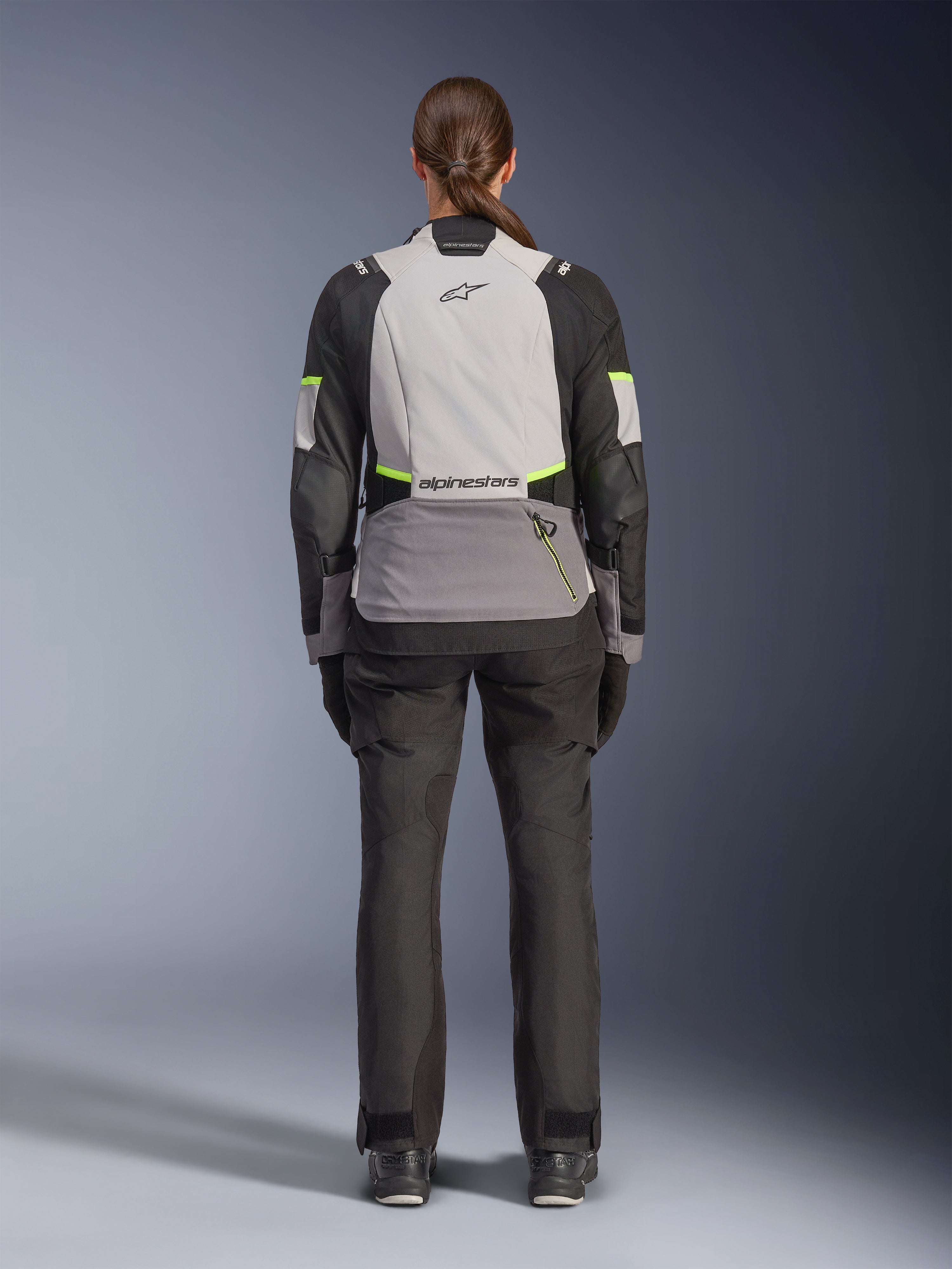 Motarde vue de dos portant la veste Alpinestars Stella Andes V4 Drystar®, Gris Glace, Gris Foncé et Noir avec accents Jaune Fluo, dotée d'une grande poche de rangement à l'arrière et du logo Alpinestars, avec un pantalon d'aventure noir assorti, équipement de touring moto toutes saisons
