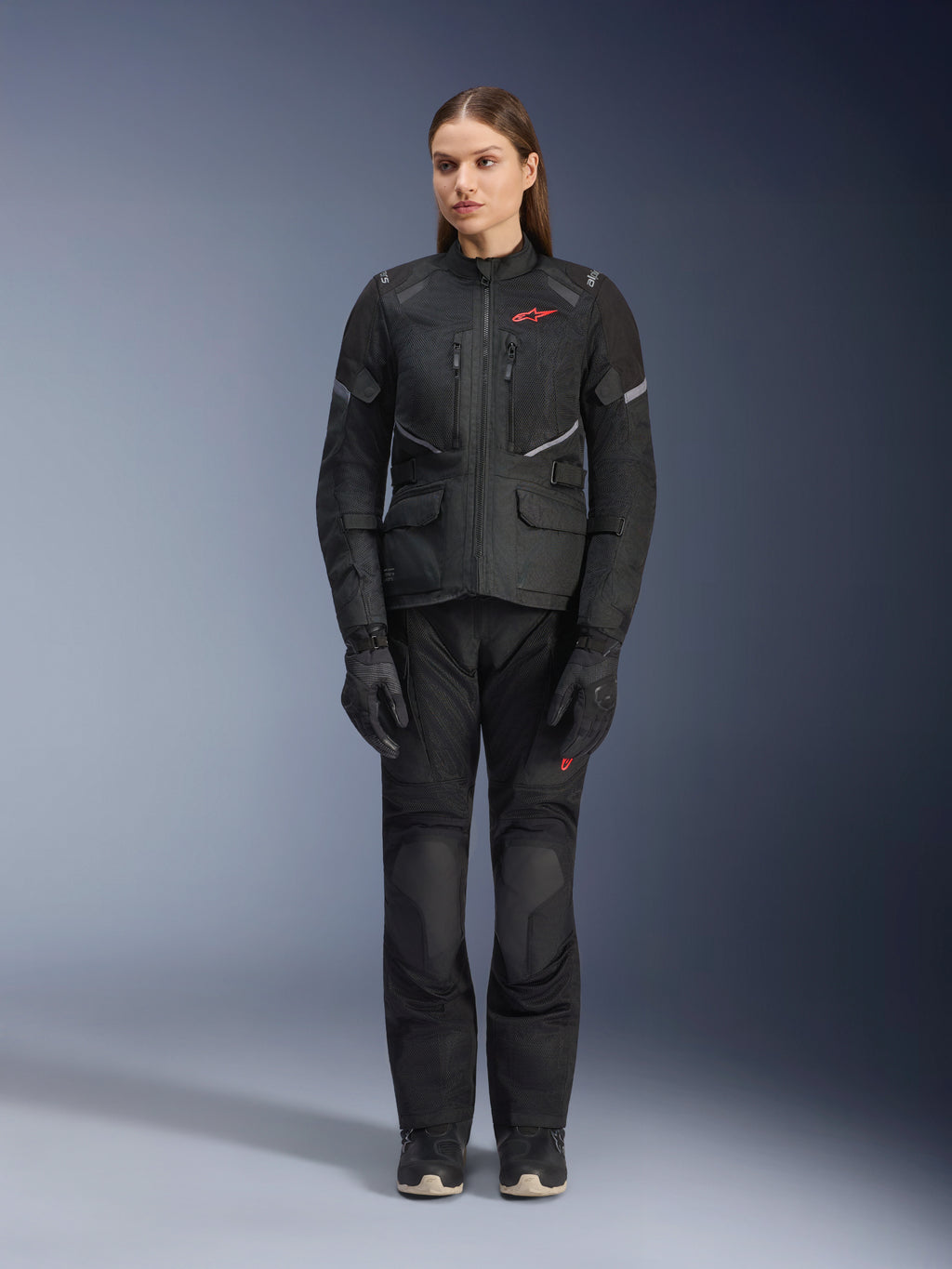 Modèle féminin portant la veste Alpinestars Stella Andes Air Drystar® noire, assortie d'un pantalon de touring et de gants noirs, avec de larges panneaux de ventilation en maille, des touches de logo rouge et des détails réfléchissants, photo de studio professionnelle pour équipement de moto pour femme.