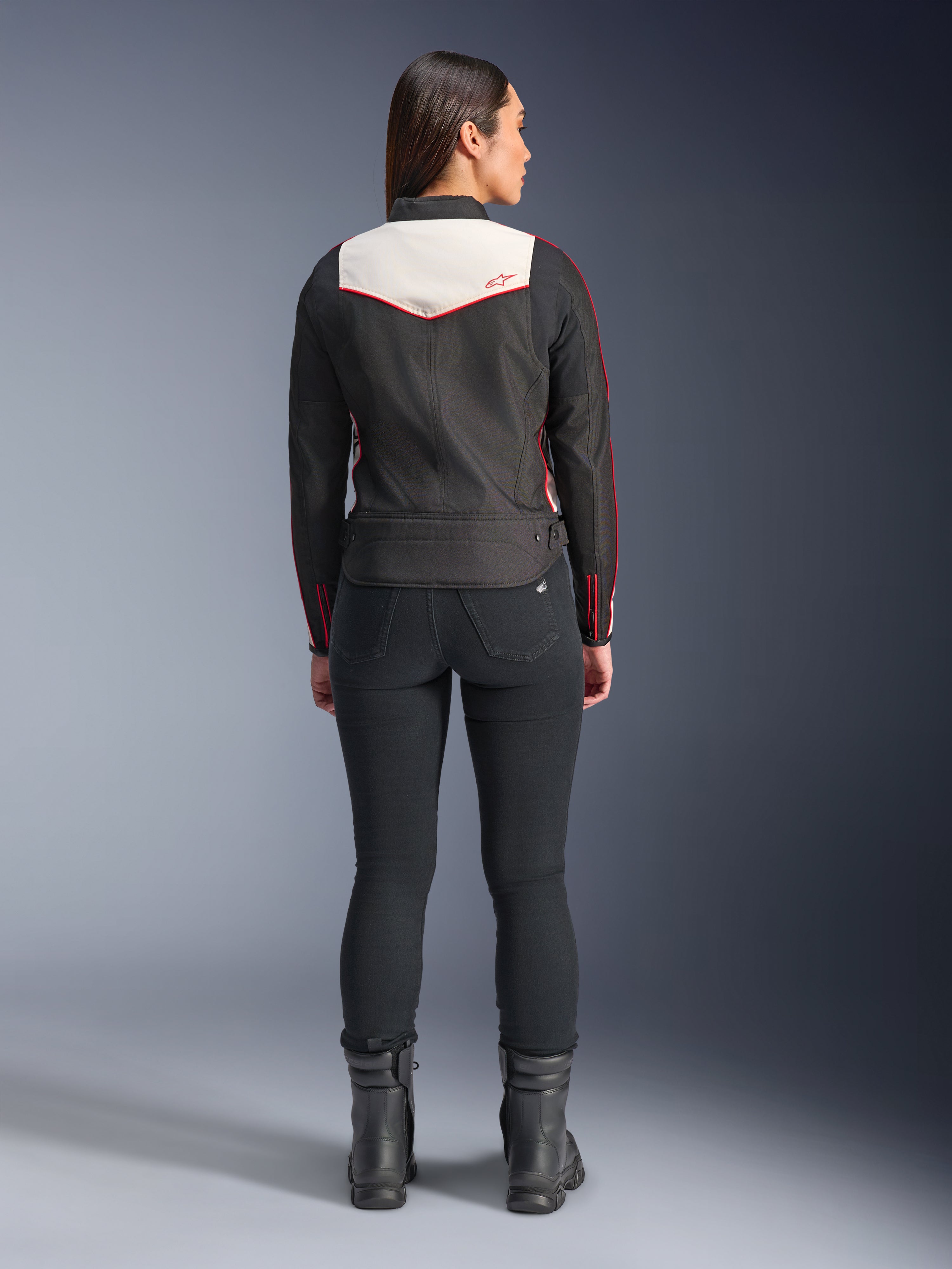 Achteraanzicht van een vrouwelijk model in de Alpinestars Stella T-Dyno WR-jas in Black Ecru Bright Red, voorzien van een opvallend crème rugpaneel en rode logo-accenten, gecombineerd met een zwarte broek en laarzen, een vintage-geïnspireerde textiele motorjas.
