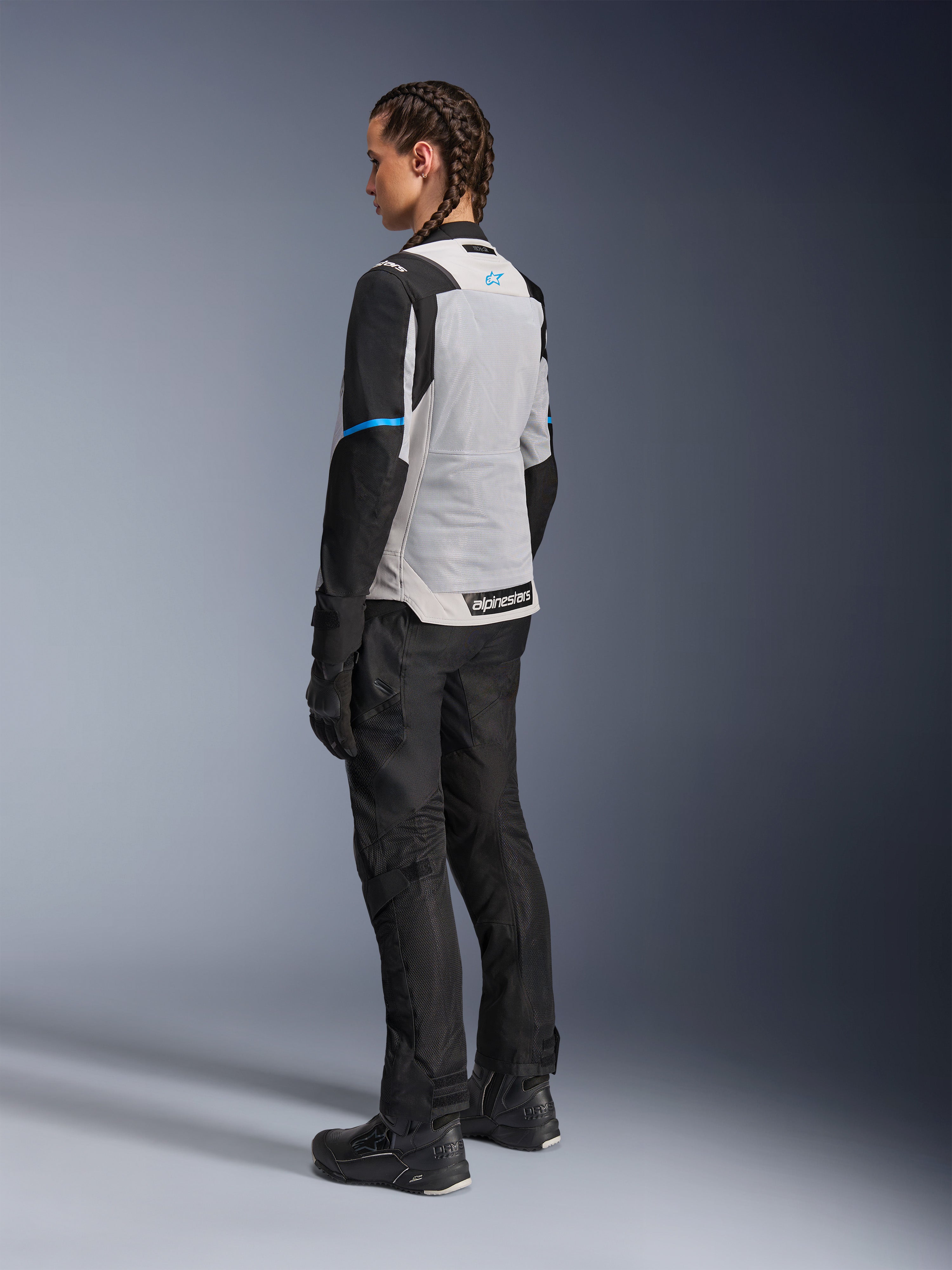 Female rider wearing the Alpinestars Stella ST-2 Air Jacket in Ice Gray and Black, rear three-quarter view highlighting the large light gray mesh back panel, black sleeves with a blue accent stripe, and a blue Alpinestars logo, paired with black riding pants, gloves, and boots for a complete adventure touring outfit