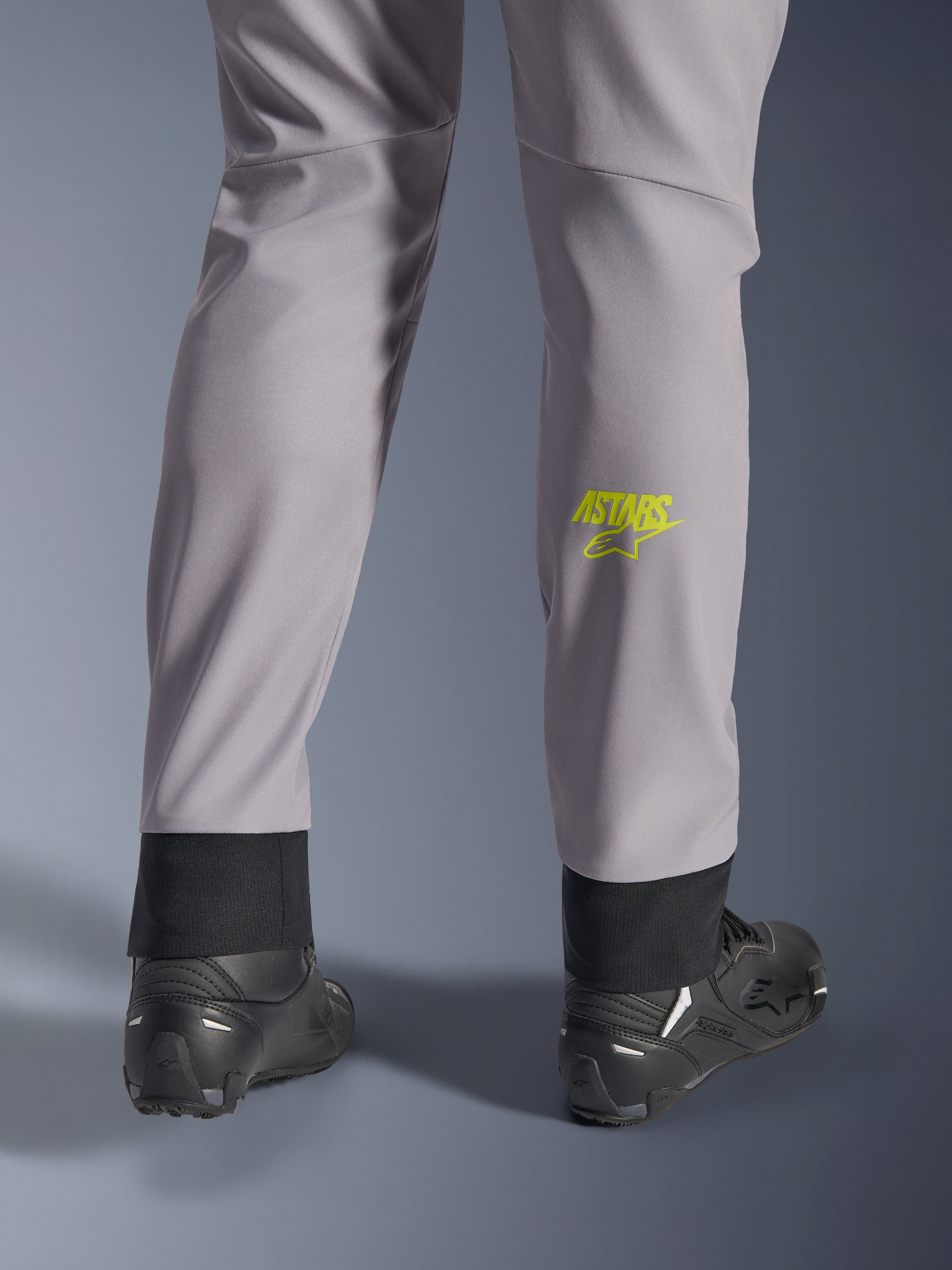 Alpinestars Aeroshell Hose, Mittelgrau mit Yellow Fluo Astars-Logo auf der Wade, Rückansicht mit schwarzen Motorradschuhen, elastischer unterer Saum und moderne, schmal zulaufende Silhouette