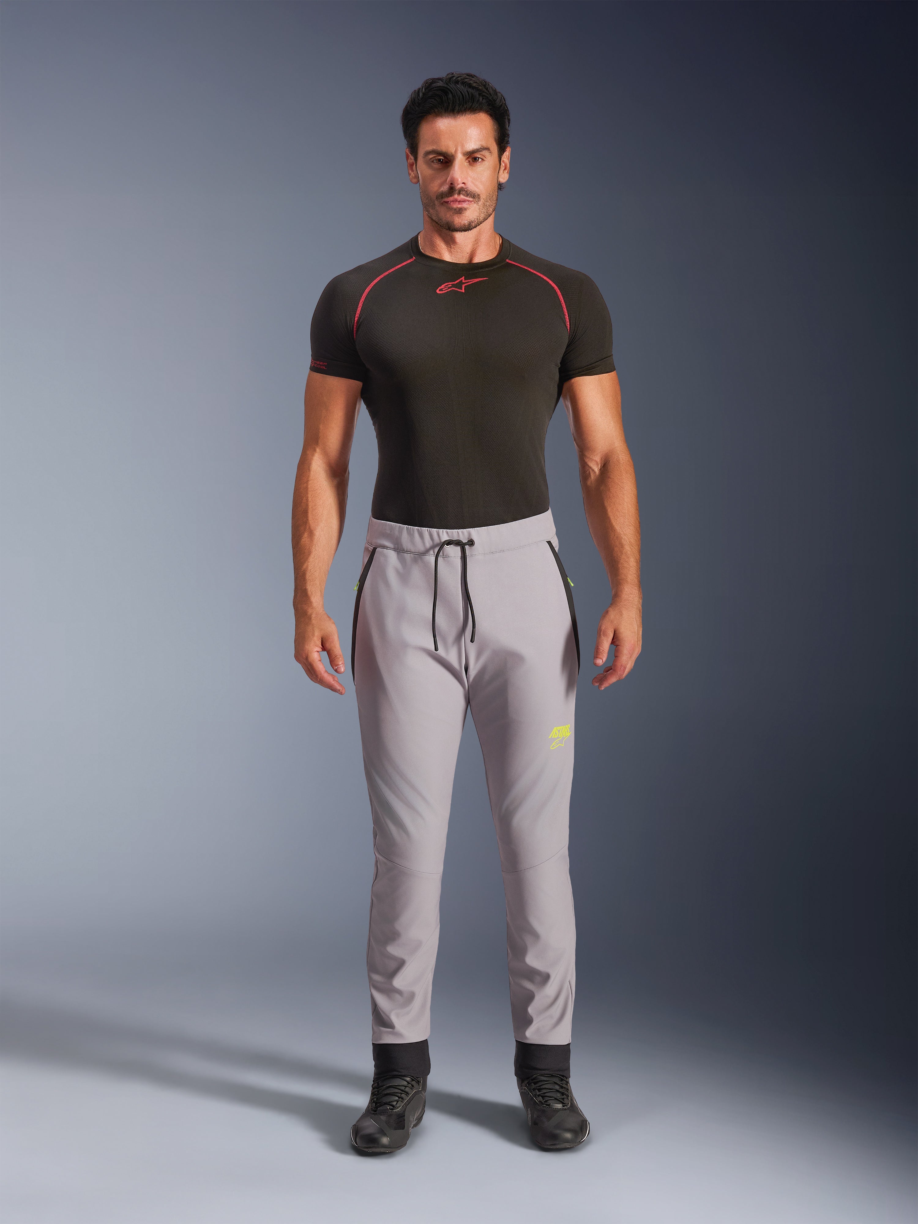 Männliches Model in Alpinestars Aeroshell Hose in Mid Gray und Yellow Fluo, Crossover-Sport-Softshellhose mit elastischem Kordelzug am Bund und Reißverschlusstaschen, kombiniert mit einem schwarzen kurzärmeligen Funktionsshirt, modernes athletisches Design für Straße und Freizeit