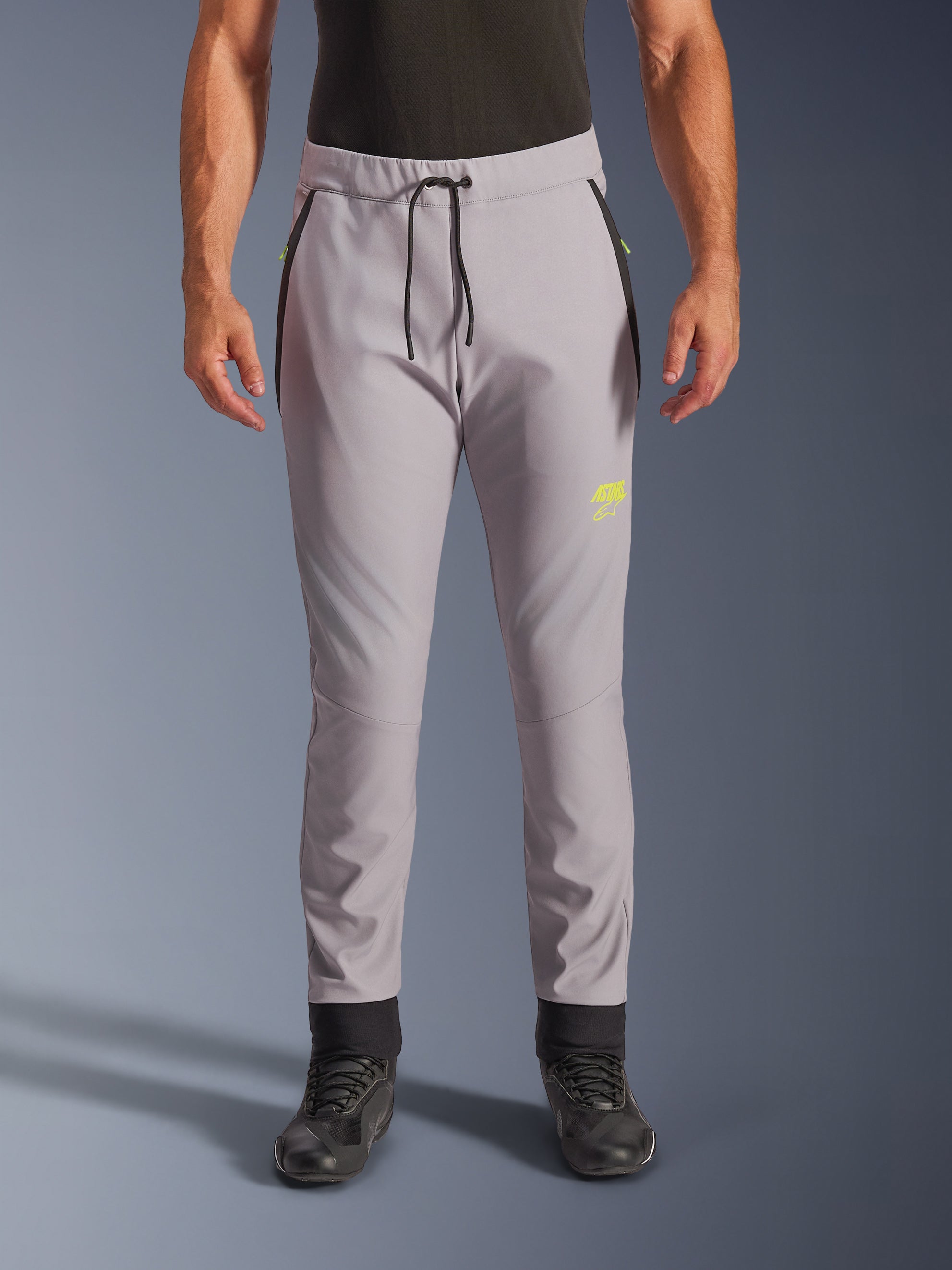 Alpinestars Aeroshell Pants, middengrijs en fluoriserend geel, crossover softshell motorbroek met een zwart trekkoord in de taille en aansluitende enkelmanchetten, geel logo op de linkerpijp, voorgevormde knieën en een sportief jogger-stijl silhouet