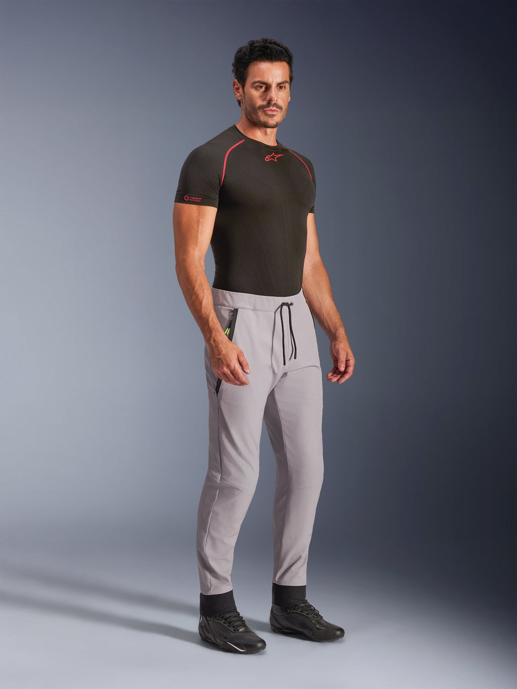 Alpinestars Aeroshell Hose, Mittelgrau mit gelben Fluo-Akzenten, Crossover-Softshell-Hose getragen von einem Model mit schwarzem Baselayer und schwarzen Schuhen, mit moderner, schmal zulaufender Passform, elastischem Bund mit schwarzem Kordelzug und Reißverschlusstaschen.