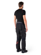 Alpinestars Hyde XT Drystar® Hose, Adventure-Tourenhose, Schwarz und Hellrot, Rückansicht eines Fahrers mit schwarzem Base-Layer und schwarzen Stiefeln, strapazierfähige Textilkonstruktion mit rotem Stern-Logo am Unterschenkel und grauem Alpinestars-Branding am Bund