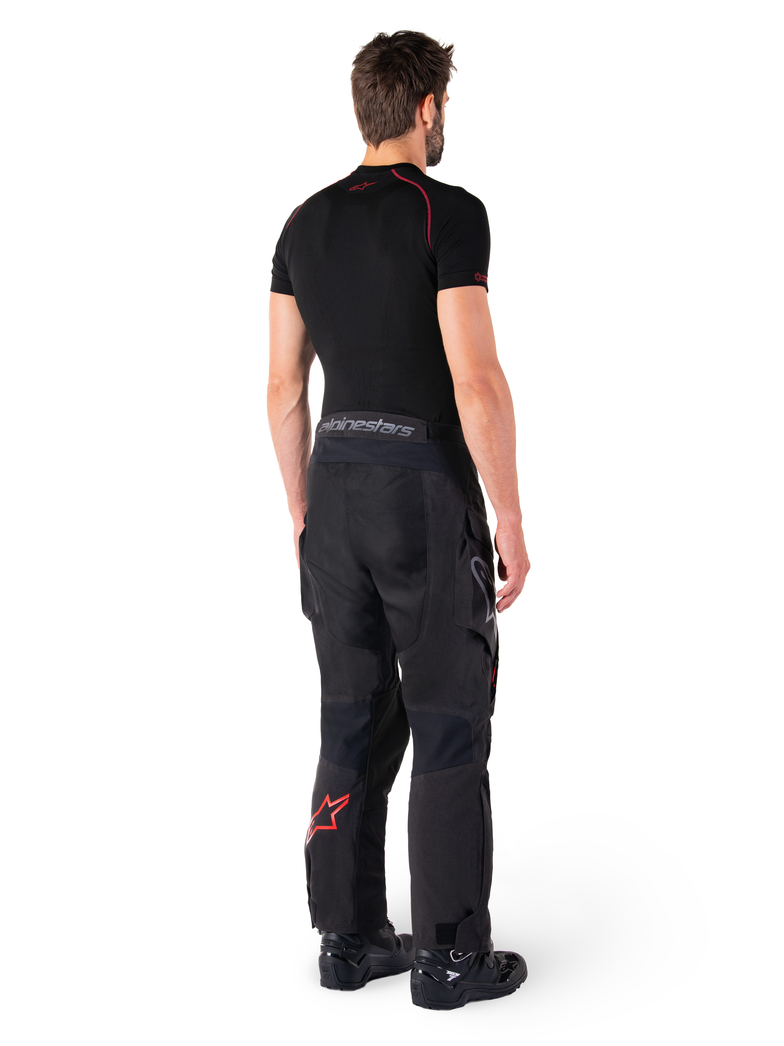 Alpinestars Hyde XT Drystar® Hose, Adventure-Tourenhose, Schwarz und Hellrot, Rückansicht eines Fahrers mit schwarzem Base-Layer und schwarzen Stiefeln, strapazierfähige Textilkonstruktion mit rotem Stern-Logo am Unterschenkel und grauem Alpinestars-Branding am Bund