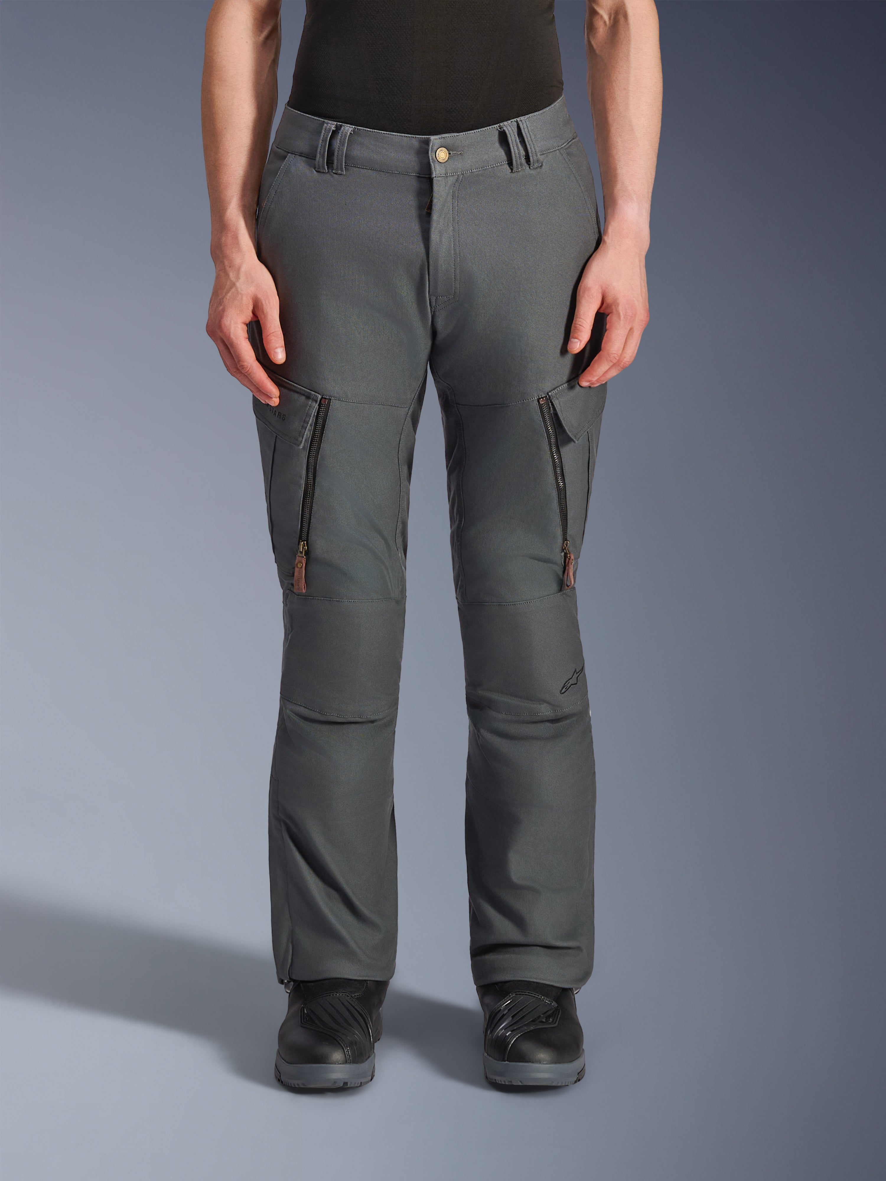 Pantalon Alpinestars Borrego Drystar®, pantalon de moto aventure, gris anthracite, doté de poches zippées verticales sur les cuisses avec tirettes en cuir, fermeture à bouton à la taille et passants de ceinture, porté par un mannequin avec des bottes de moto noires