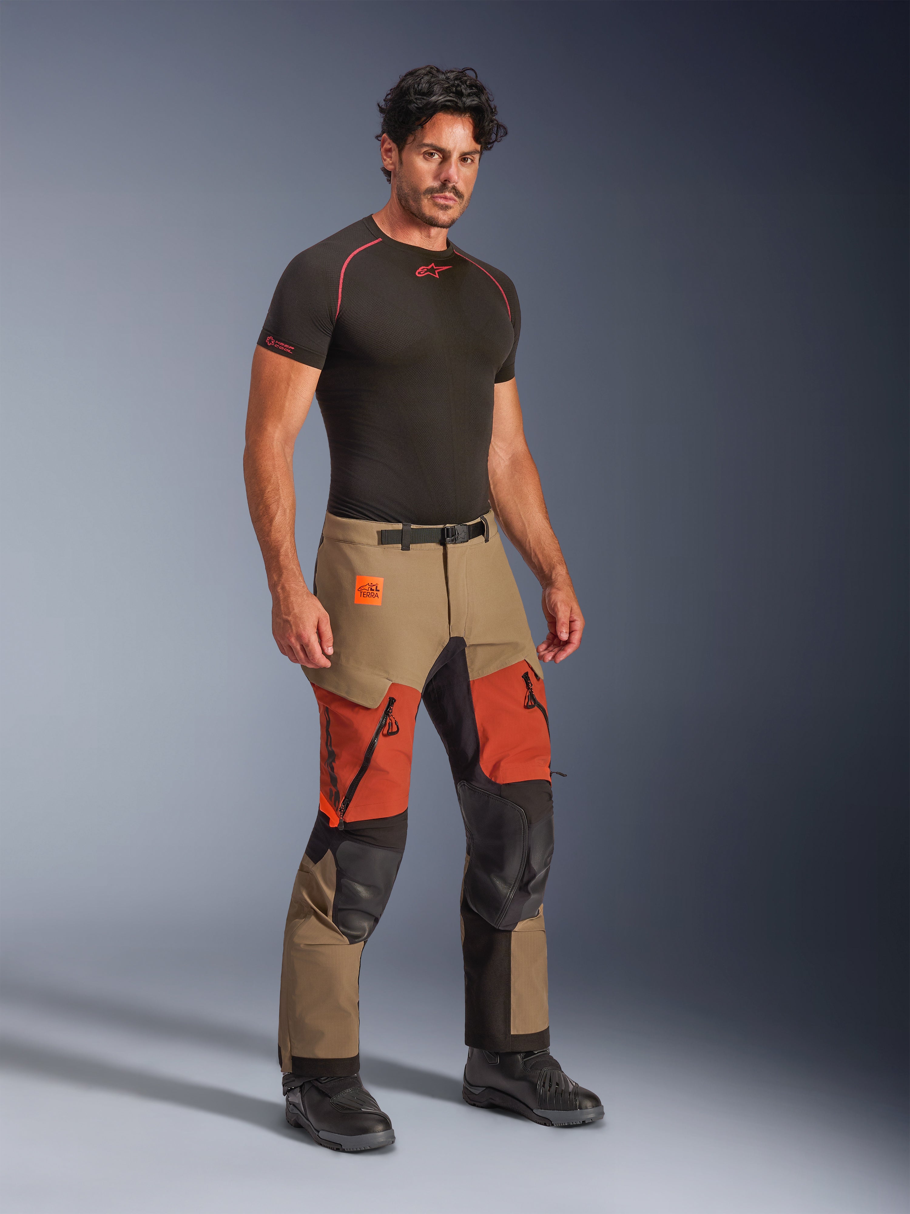 Alpinestars Halo Pro Drystar® XF Gelamineerde Broek, adventure motorbroek, Walnut Rust en zwart, gedragen door een model met een zwarte compressie-onderlaag en zwarte laarzen, voorzien van versterkte kniepanelen, oranje dijventilatieopeningen met rits en Terra-branding, technische all-terrain rijuitrusting