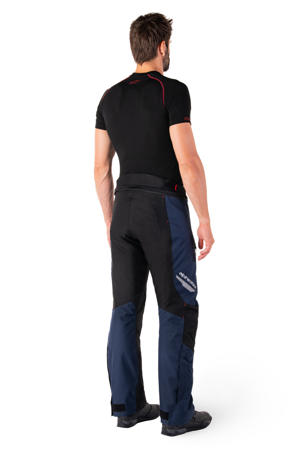 Pantalon imperméable Alpinestars St-1, pantalon touring aventure, bleu foncé, noir et rouge vif, vue arrière latérale, porté avec une couche de base noire à manches courtes et des bottes de moto noires, doté d'un design épuré avec marquage réfléchissant et accents rouges