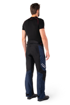 Pantalon imperméable Alpinestars St-1, pantalon touring aventure, bleu foncé, noir et rouge vif, vue arrière latérale, porté avec une couche de base noire à manches courtes et des bottes de moto noires, doté d'un design épuré avec marquage réfléchissant et accents rouges