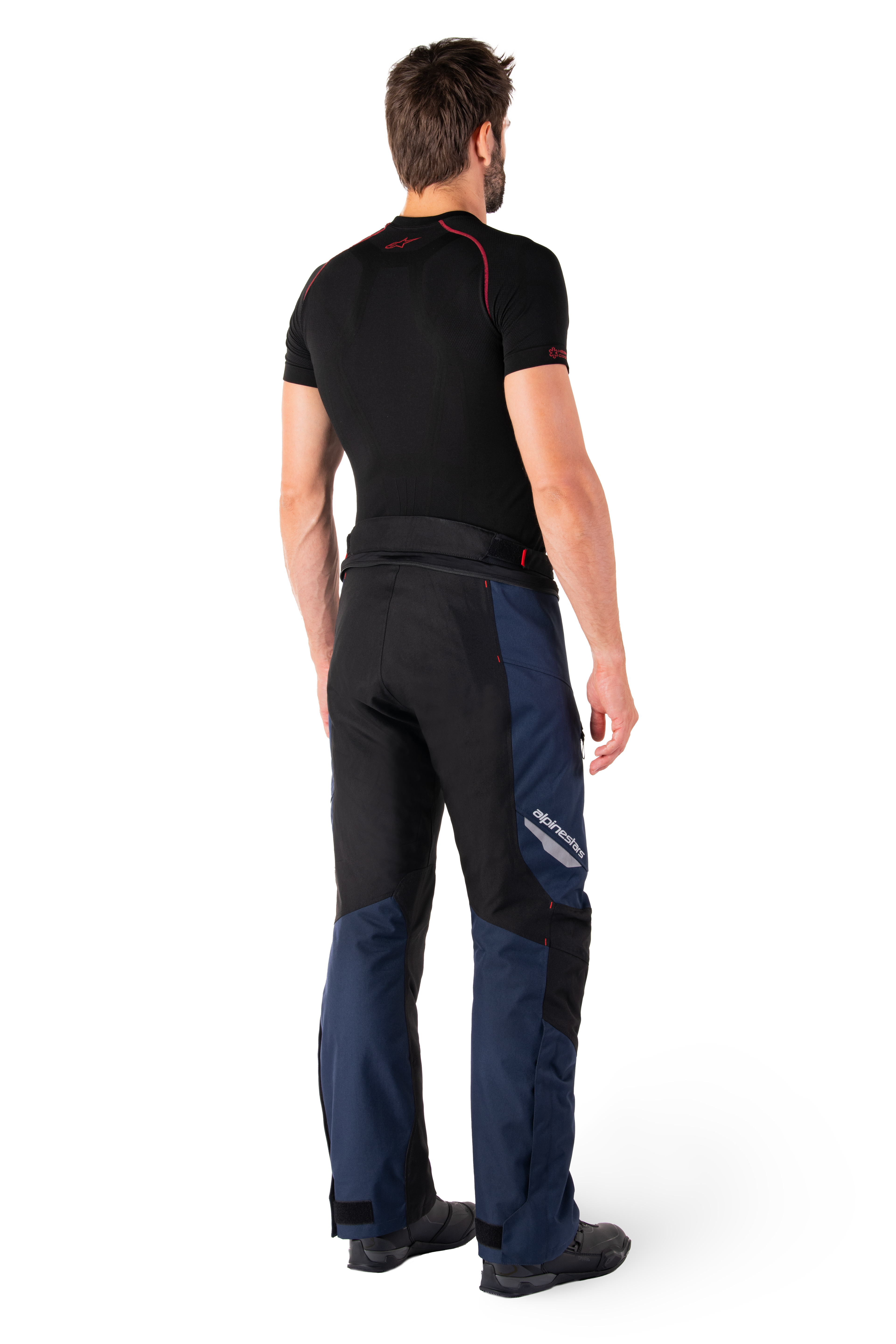Pantalon imperméable Alpinestars St-1, pantalon touring aventure, bleu foncé, noir et rouge vif, vue arrière latérale, porté avec une couche de base noire à manches courtes et des bottes de moto noires, doté d'un design épuré avec marquage réfléchissant et accents rouges